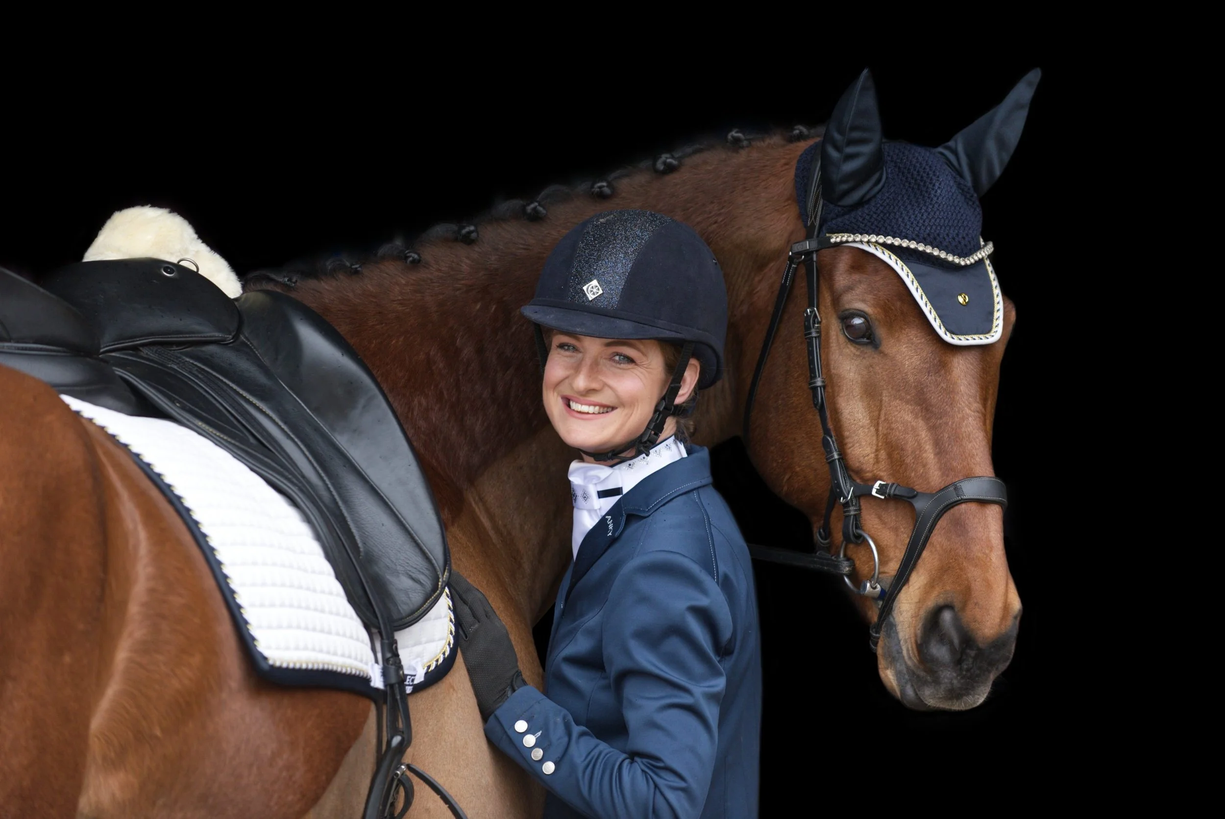 A smiling woman in equestrian attire with a black riding helmet, standing next to a brown horse with a decorated bridle and a blue ear net, against a black background.
