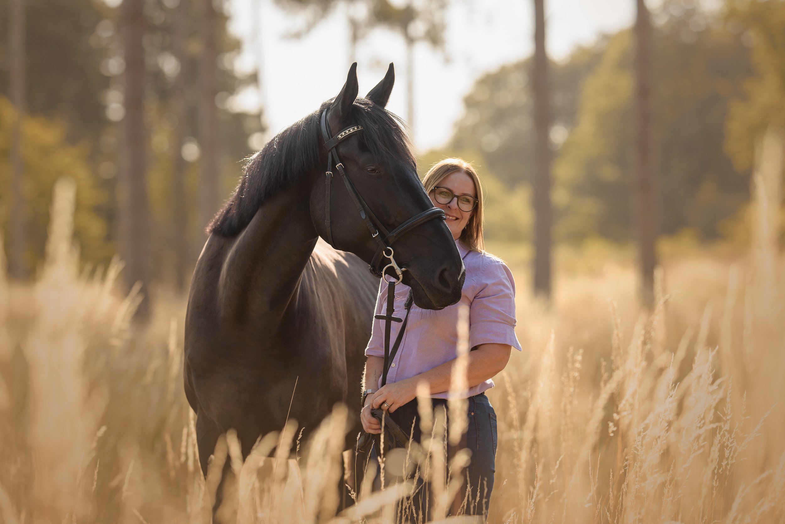 A woman with glasses smiling as she stands next to a dark horse in a field of tall, golden grass with a forest of trees in the background during sunset.