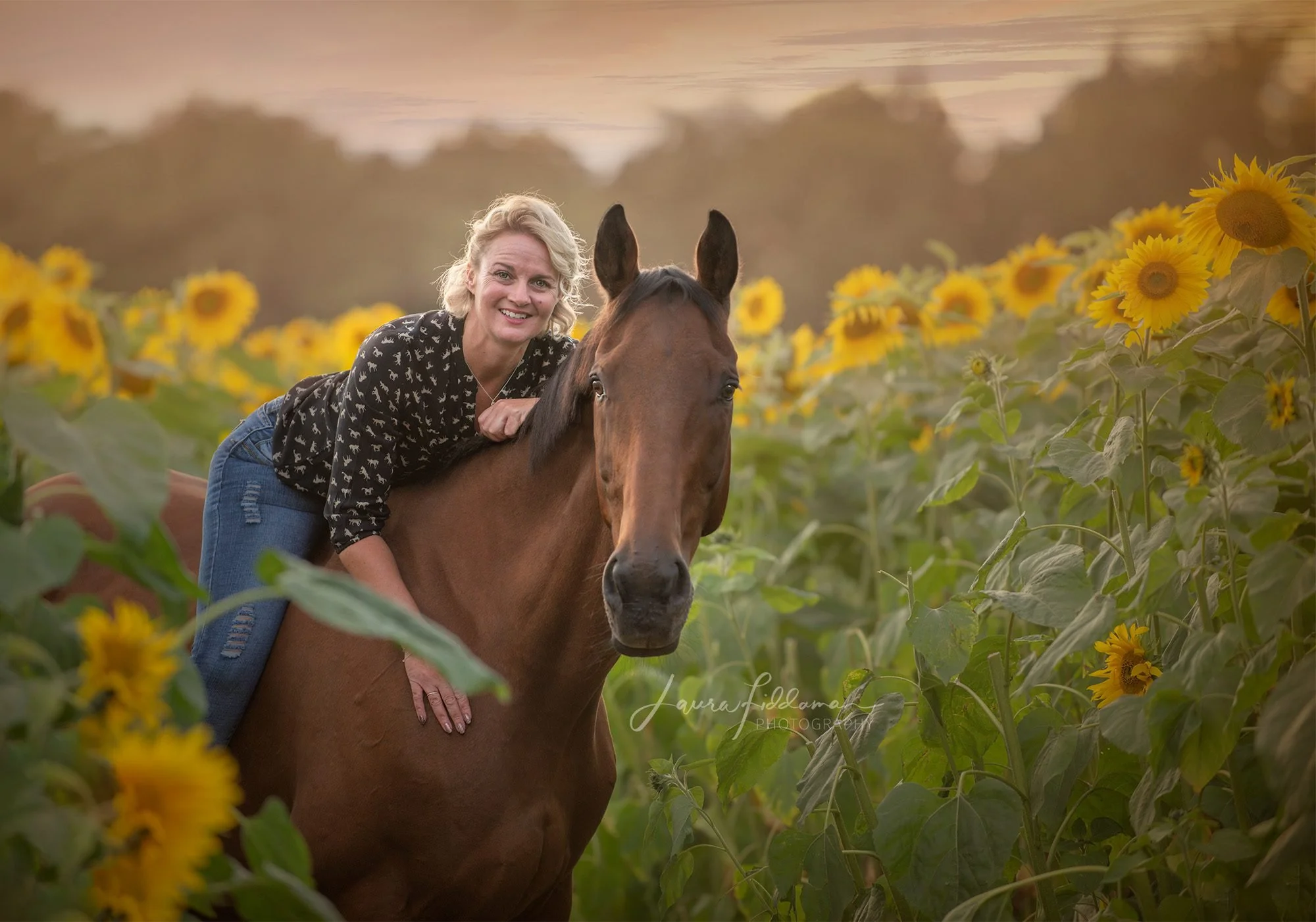A smiling woman with blonde hair wearing a black blouse with white dog patterns, riding a brown horse through a sunflower field during sunset.
