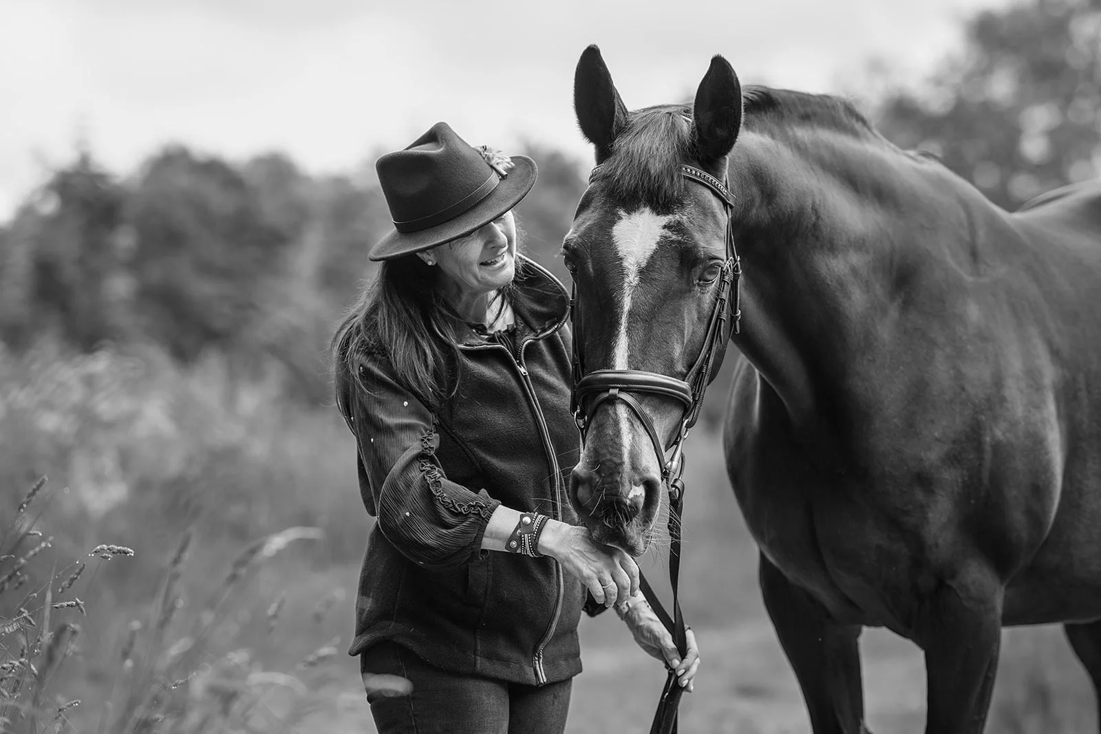 A woman with long dark hair wearing a hat and a jacket interacts with a horse in an outdoor setting. The woman is smiling and touching the horse's face while the horse looks directly at the camera. The background contains blurred trees and grass.