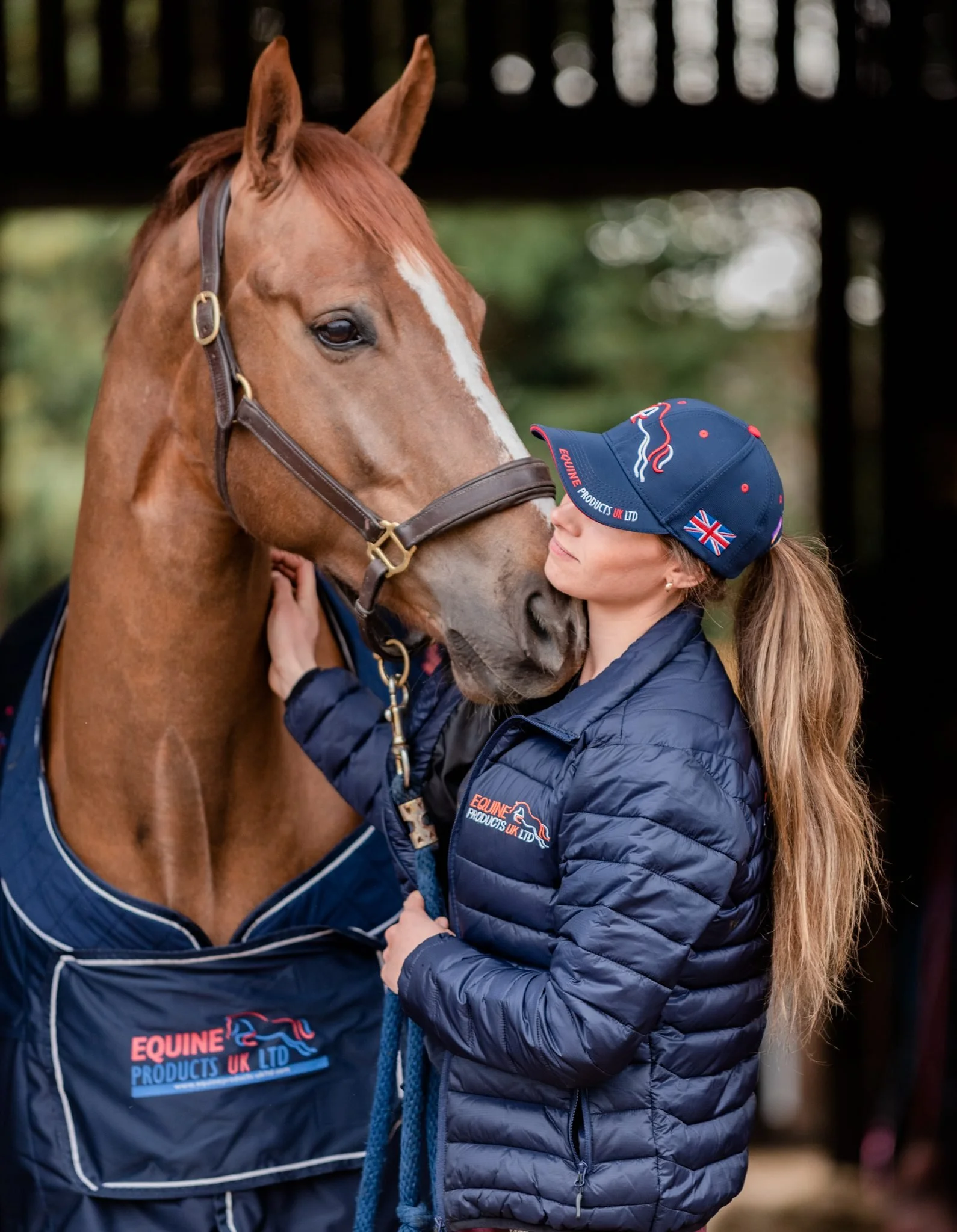 A woman and a horse touching noses outside, both wearing equine apparel, with the woman wearing a blue cap and jacket with the logo 'Equine Products UK Ltd'.