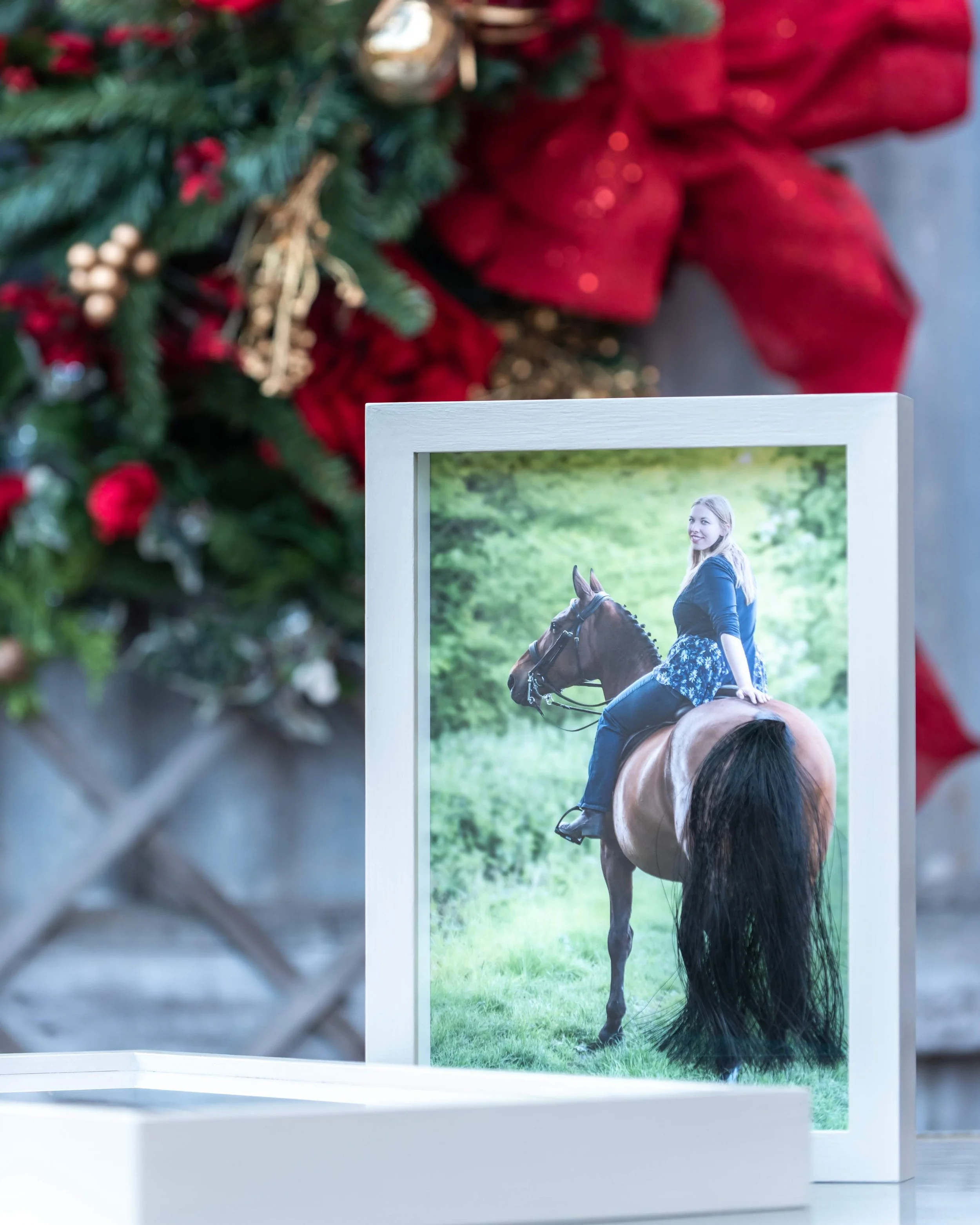 A framed photograph of a woman riding a horse outdoors, with green foliage in the background, placed on a white surface. The frame has horse tail hair inlaid as a keepsake frame.