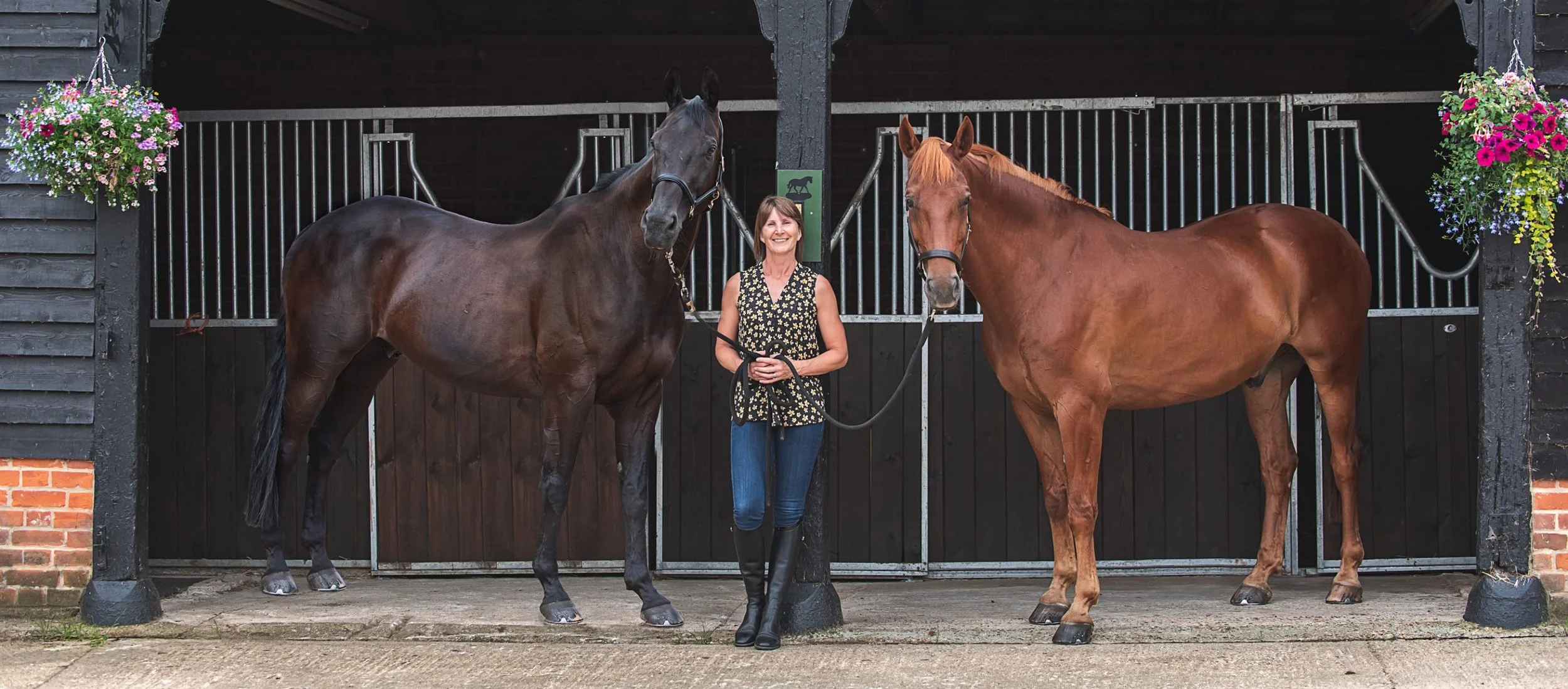 A woman standing between two horses in front of a stable, holding their reins, with hanging flower baskets on each side at Beckingham Hall.