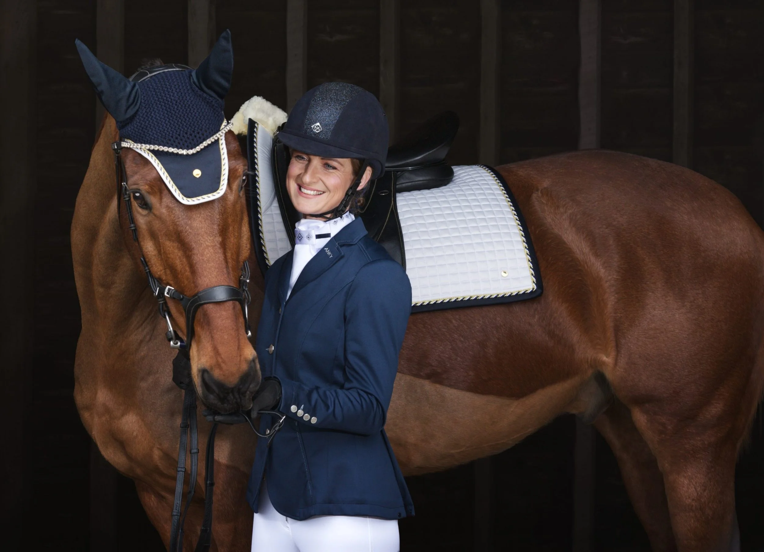 A woman smiling in tidy dressage gear hugging a bay horse with a white saddle pad in a stable.