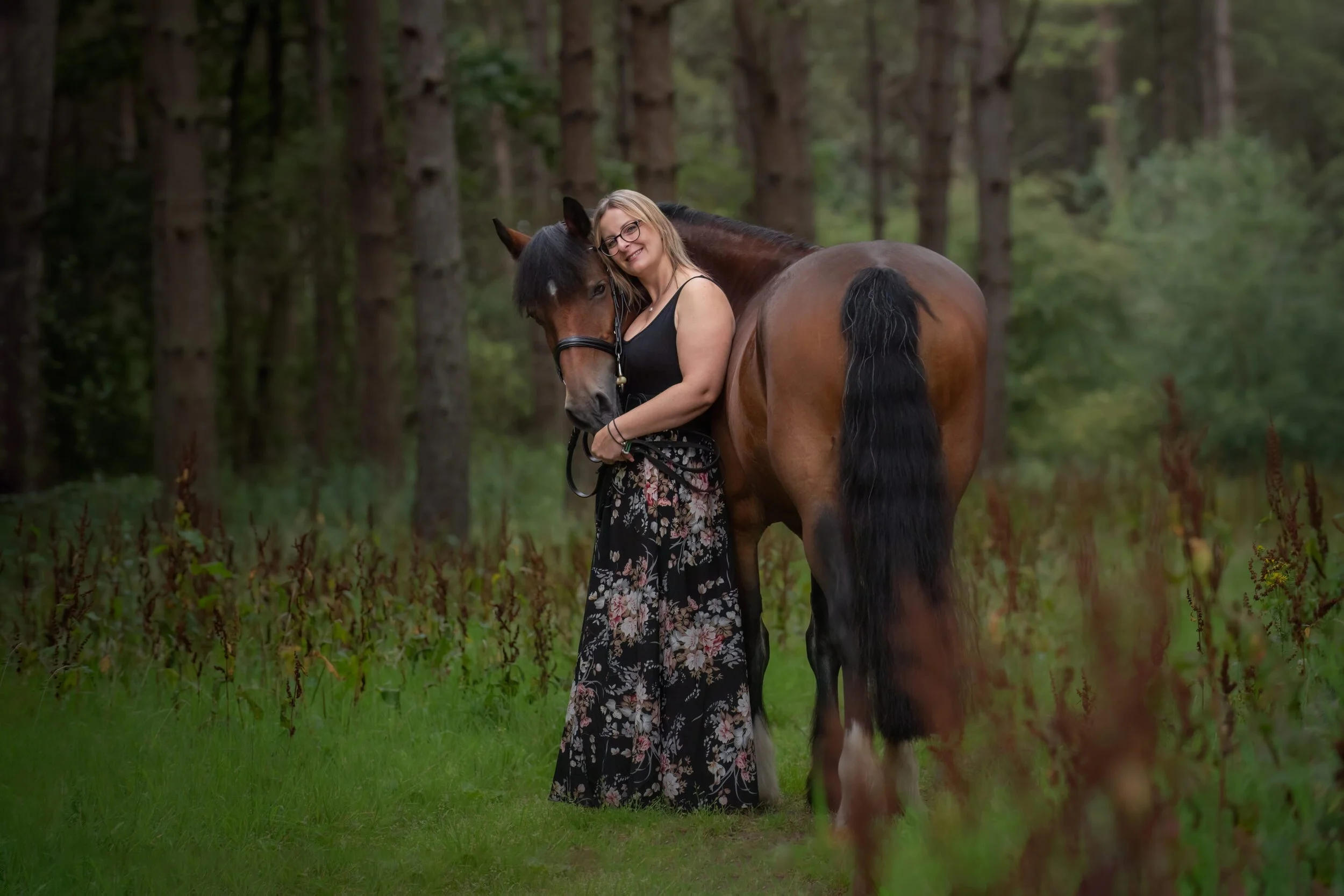 A woman with glasses, blonde hair, wearing a black tank top and floral skirt, is hugging a bay horse with a black mane in a wooded area with green grass and trees.