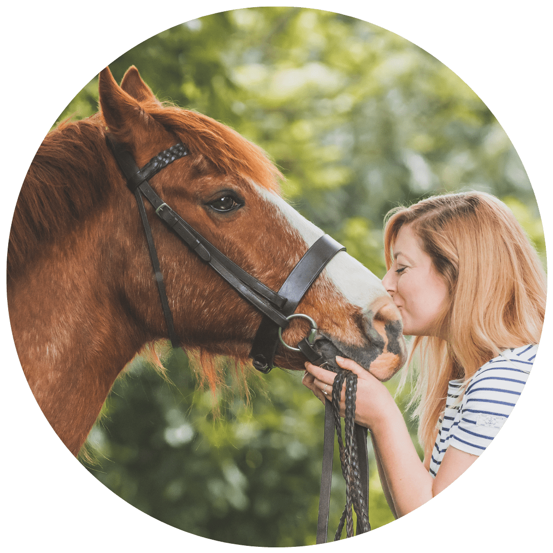 A woman with red hair shows affection to a brown and white horse with a black bridle in a green outdoor setting.