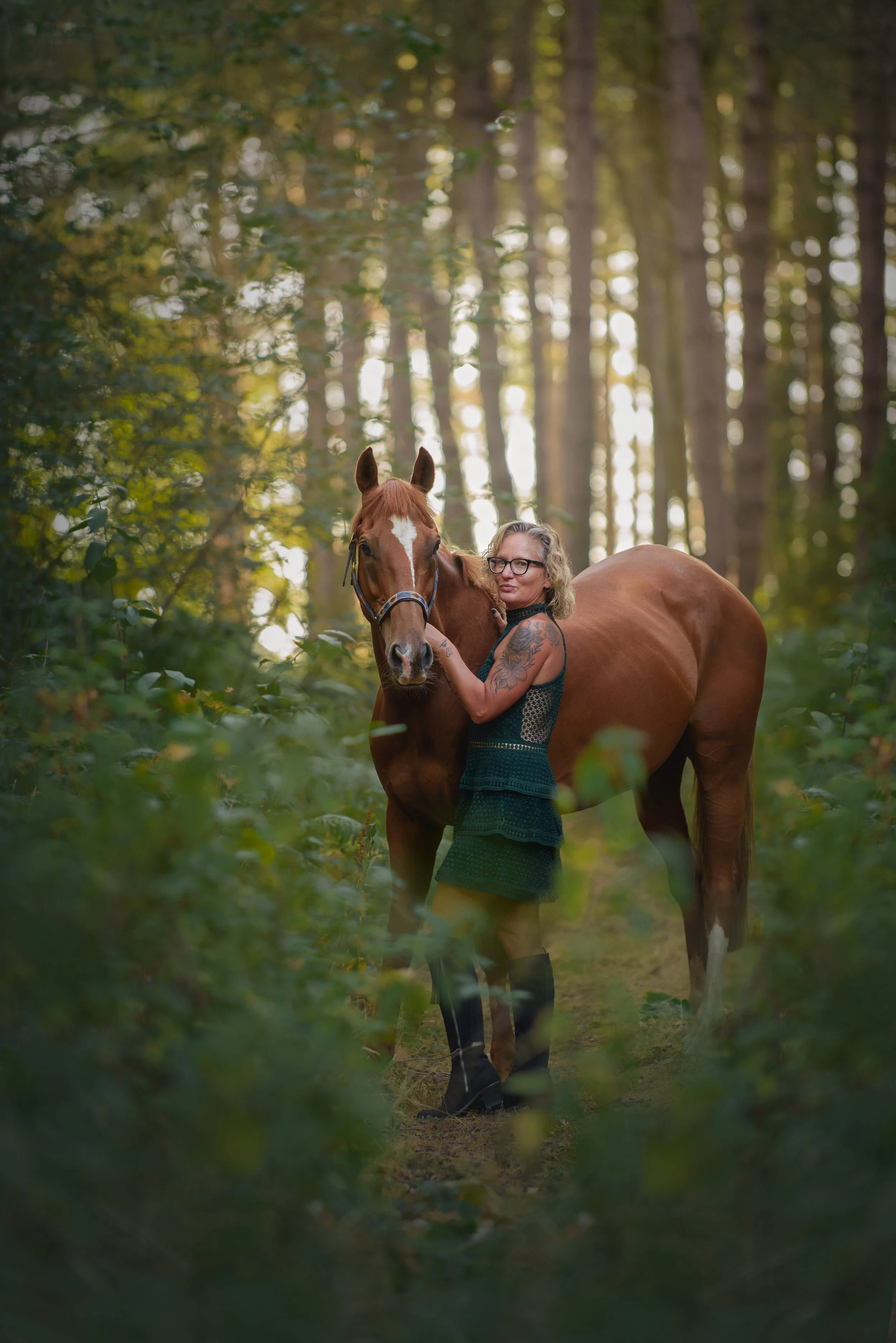 A woman with glasses and tattoos is standing next to a brown horse with a white star on its forehead in a forest clearing, surrounded by tall trees and green foliage, during daytime.