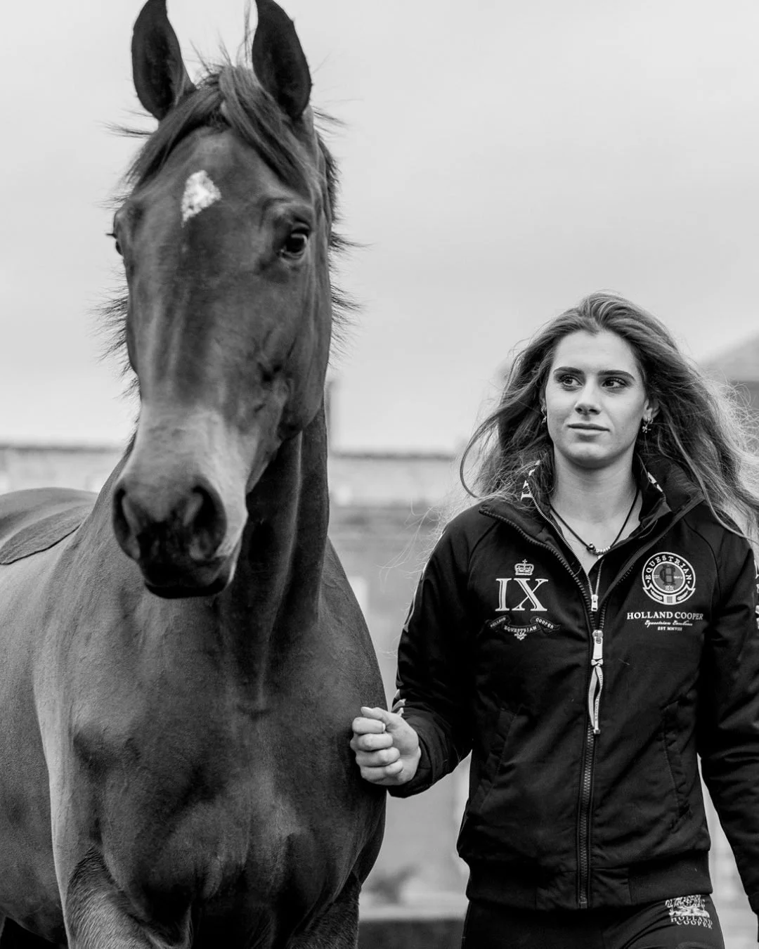 Black and white photo of a young woman standing next to a horse outdoors. The woman has long hair and is wearing a jacket with logos, while the horse has a white spot on its forehead.