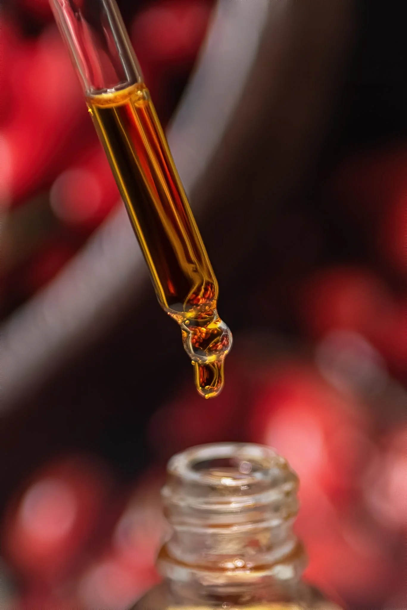 Close-up of a glass dropper with amber-colored liquid, held above a glass container, with a blurred background of red and dark hues.