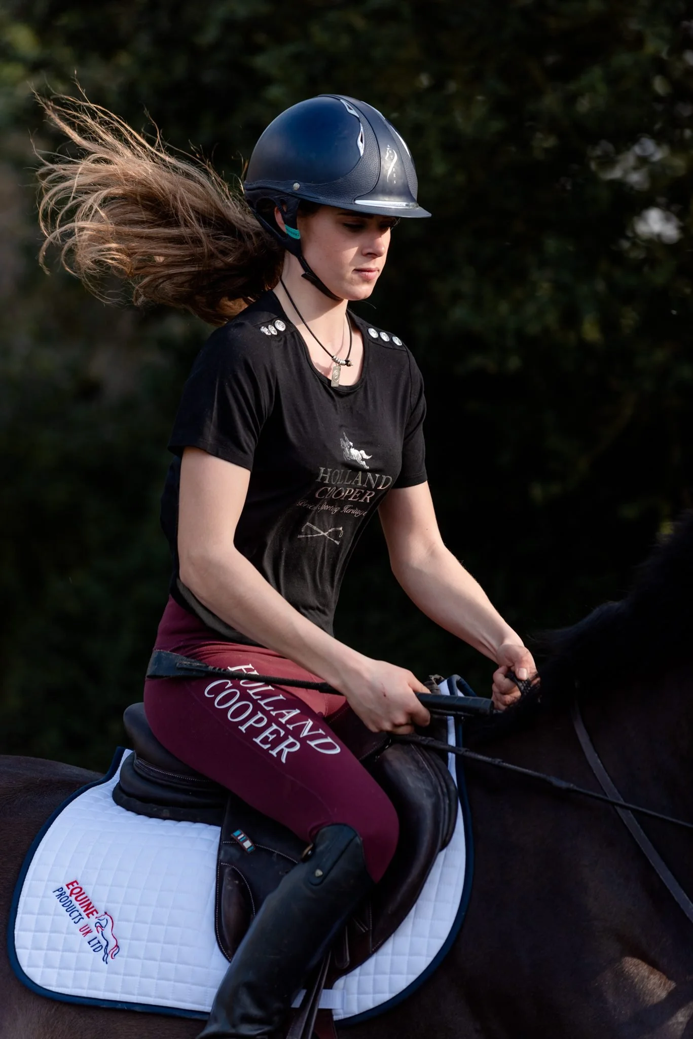 A young woman wearing a black riding helmet and maroon riding pants is riding a horse in a natural outdoor setting with trees in the background. She is dressed in equestrian Holland Cooper attire, holding the reins, and concentrating.