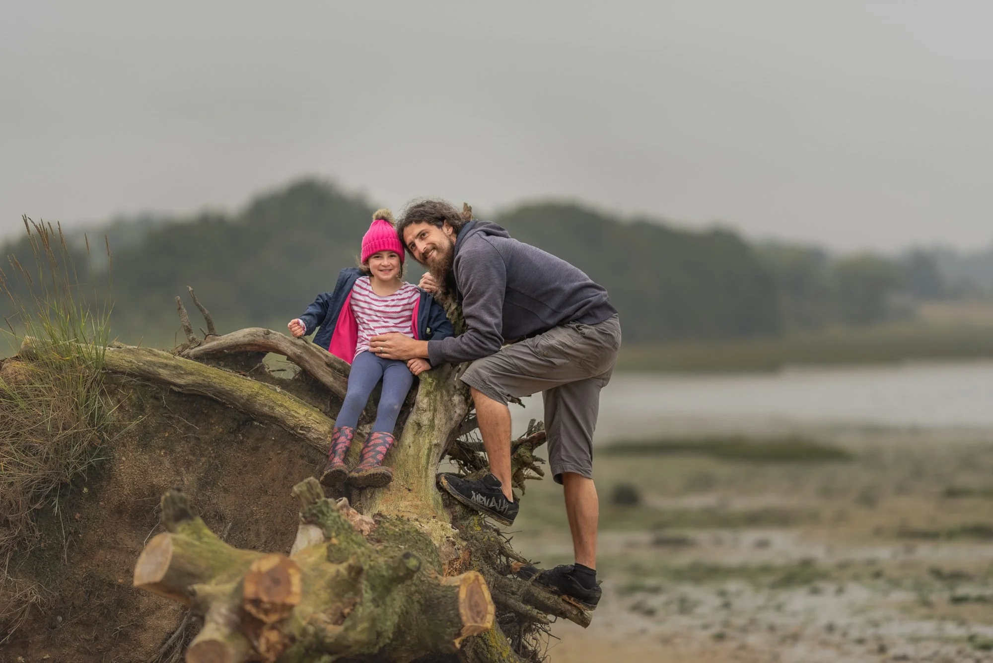 A dad and his daughter sitting on a fallen tree trunk in a marshland, smiling at the camera under cloudy skies.