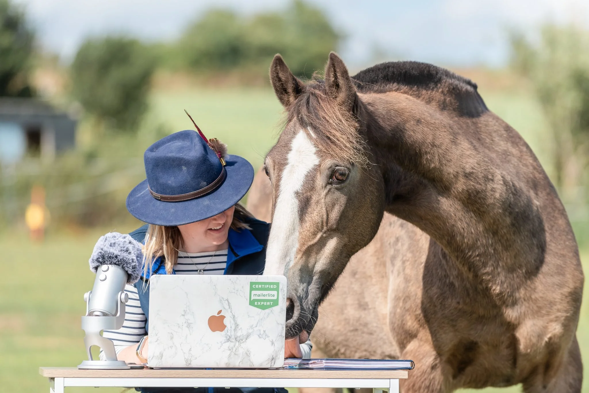 A woman in a striped shirt and a blue jacket, wearing a blue hat with a feather, is standing at a table outdoors with a laptop that has a marble pattern, a microscope, and a sign that reads "Certified MailerLite Expert." She is smiling and leaning he