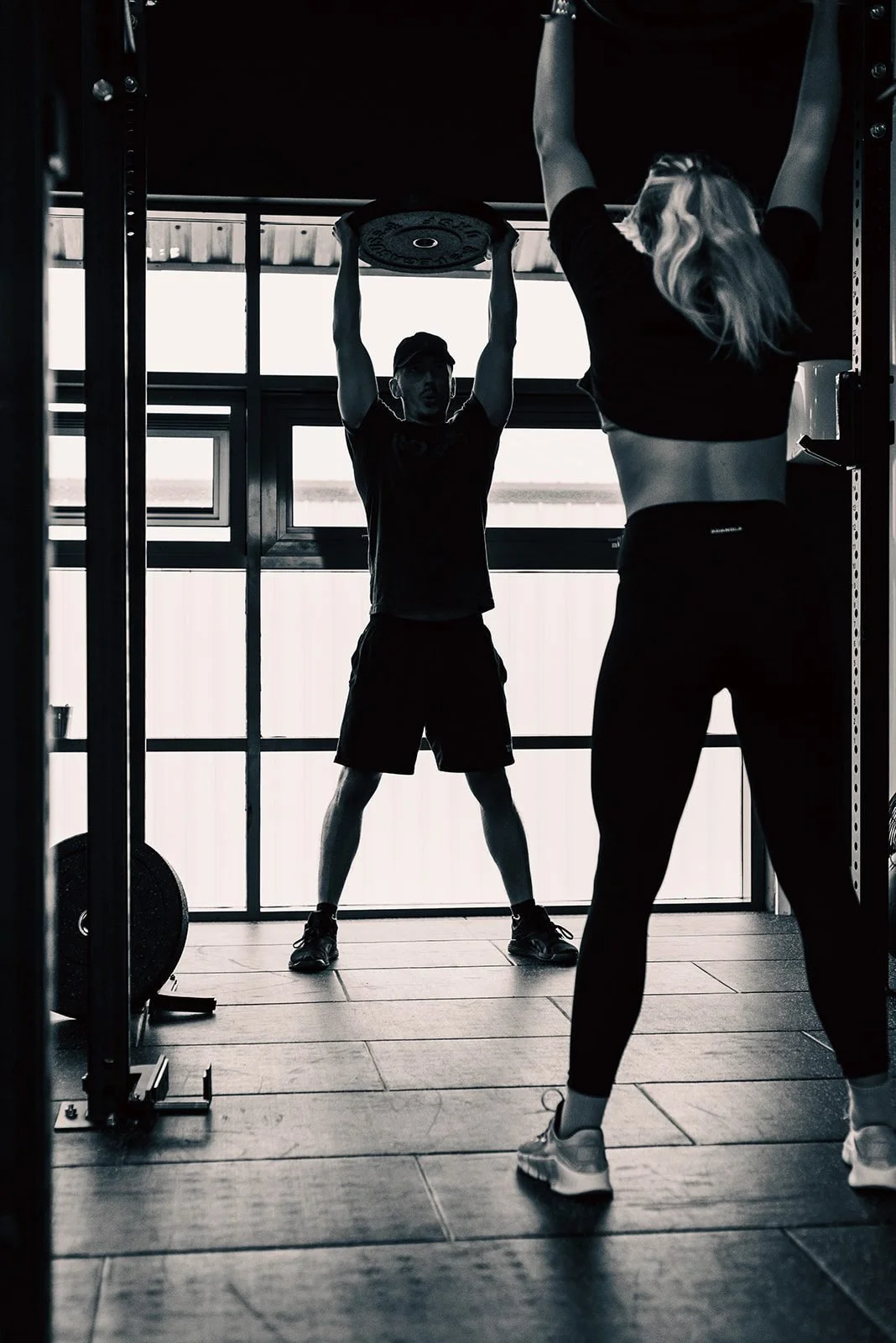 Two people working out in the Claydon Crossfit gym. One person is lifting a weight above their head while the other is standing in front, facing away, with arms raised. The gym has large windows in the background, and the photo is in black and white.