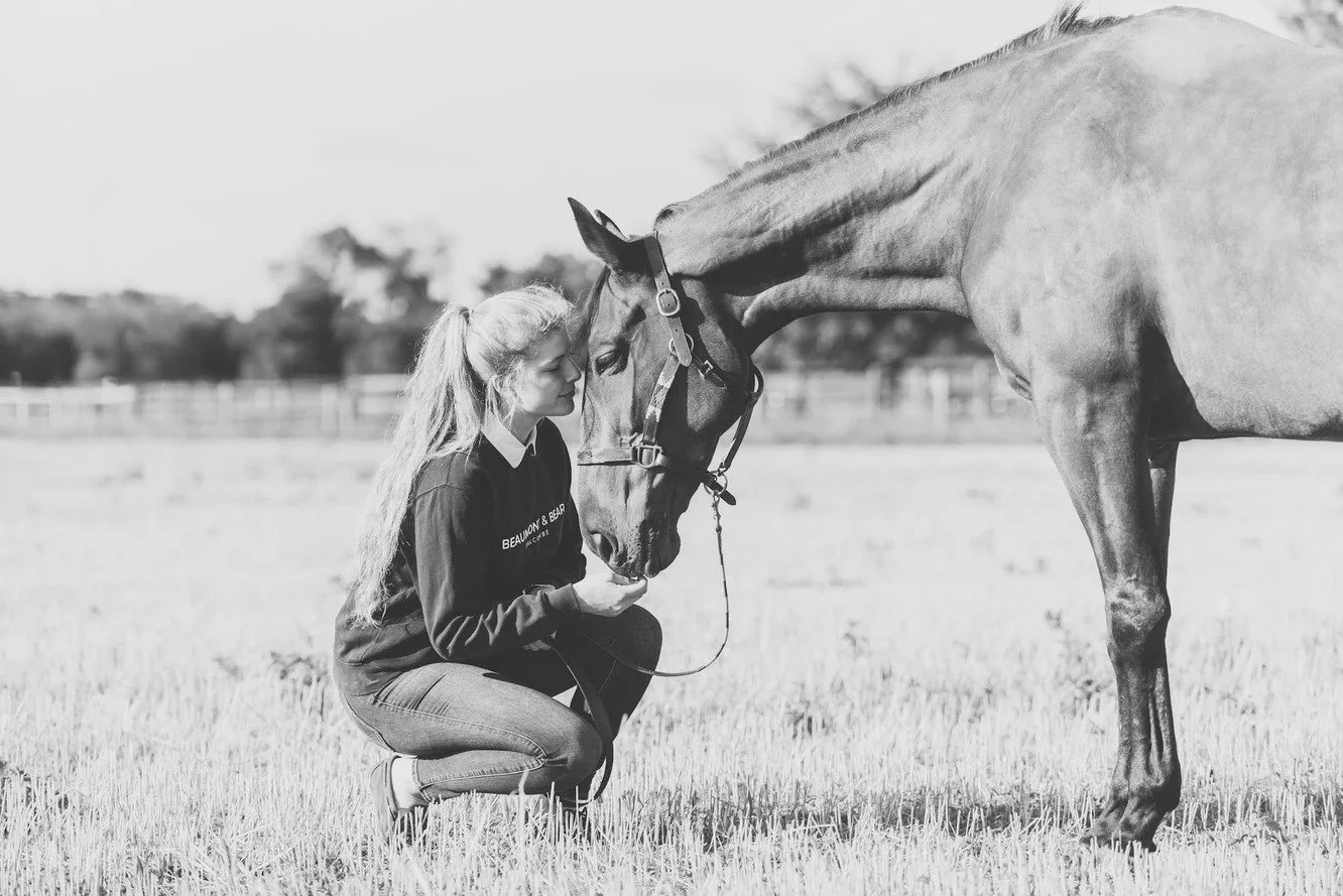 A woman kneeling in a grassy field gently touching noses with a horse. The woman has long hair tied back and is wearing a dark sweatshirt and jeans. The horse is wearing a halter. In the background, there are fences and trees, and the image is in bla