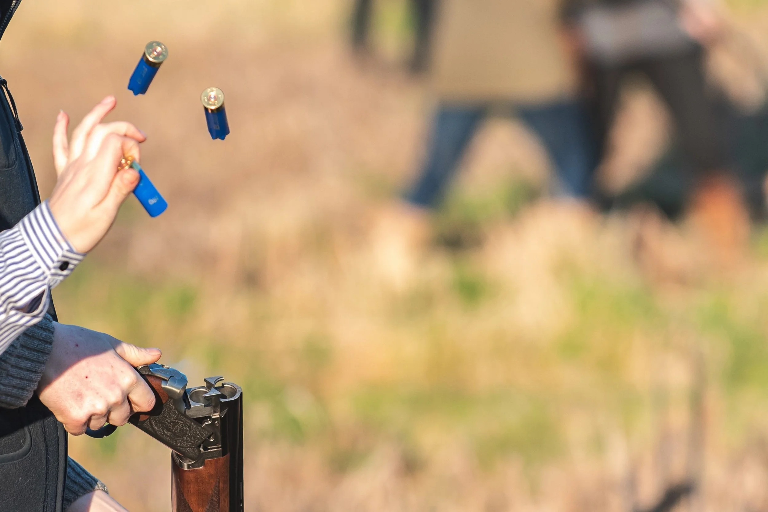 A person holding a shotgun with a hand and part of their body visible, with a blurred outdoor background.