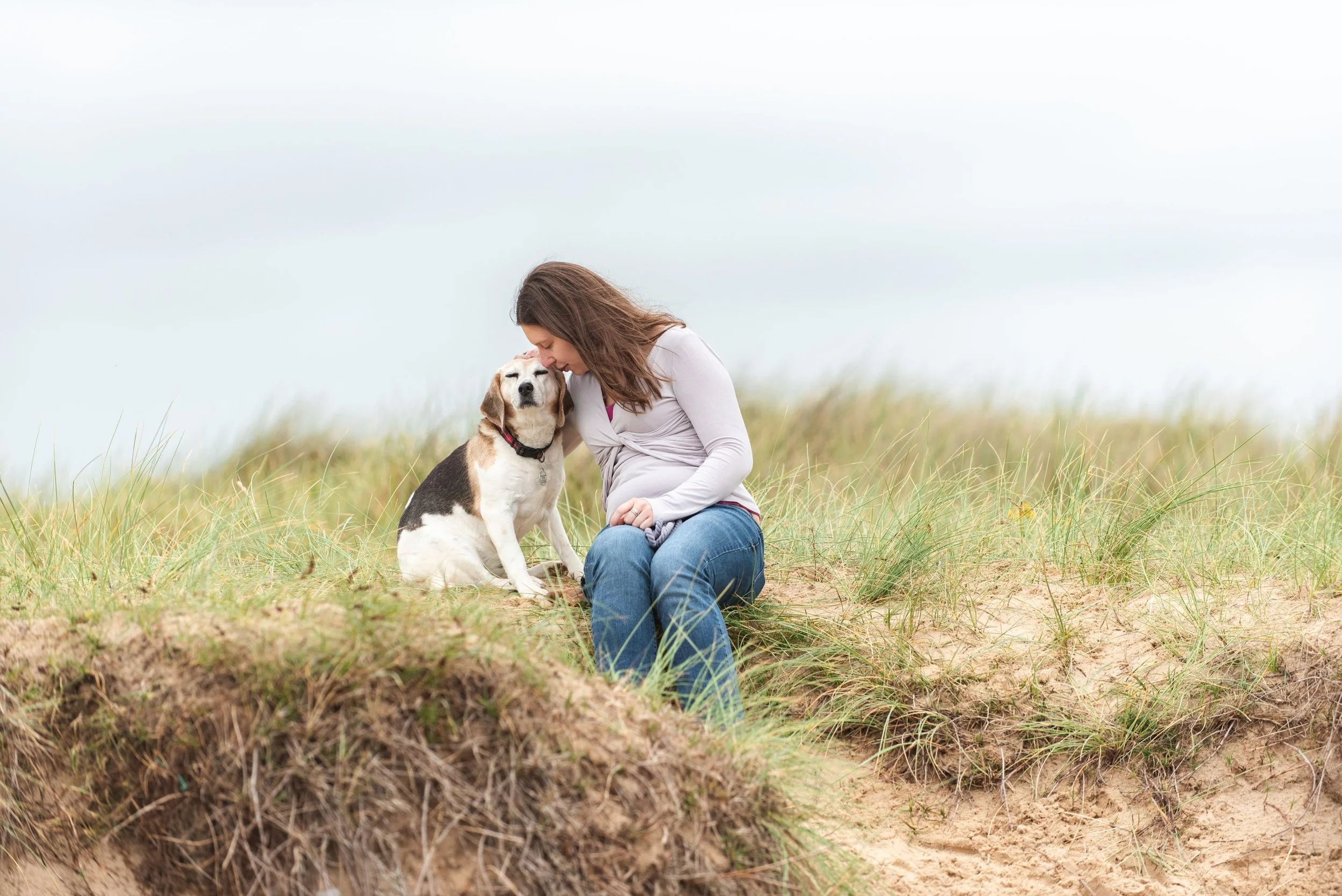 A woman with long brown hair sitting on grass, leaning her forehead against a beagle, at Walberswick beach with overcast sky.