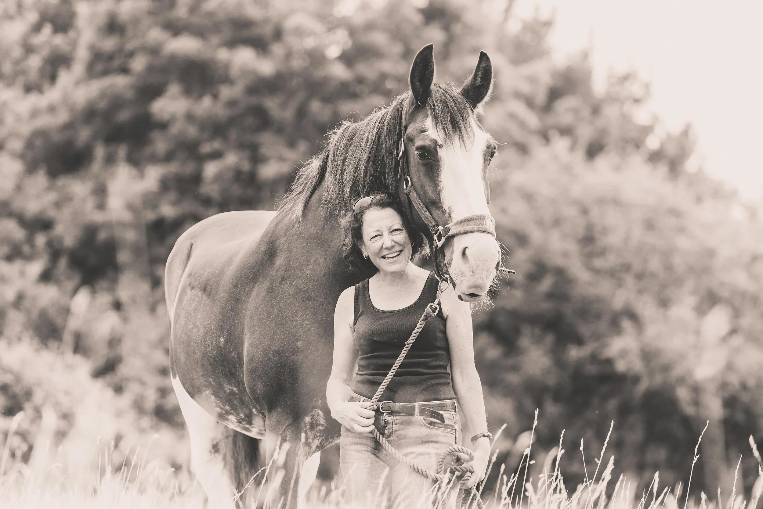 A woman smiling and holding a lead rope while standing beside a large horse in an outdoor setting with trees in the background.