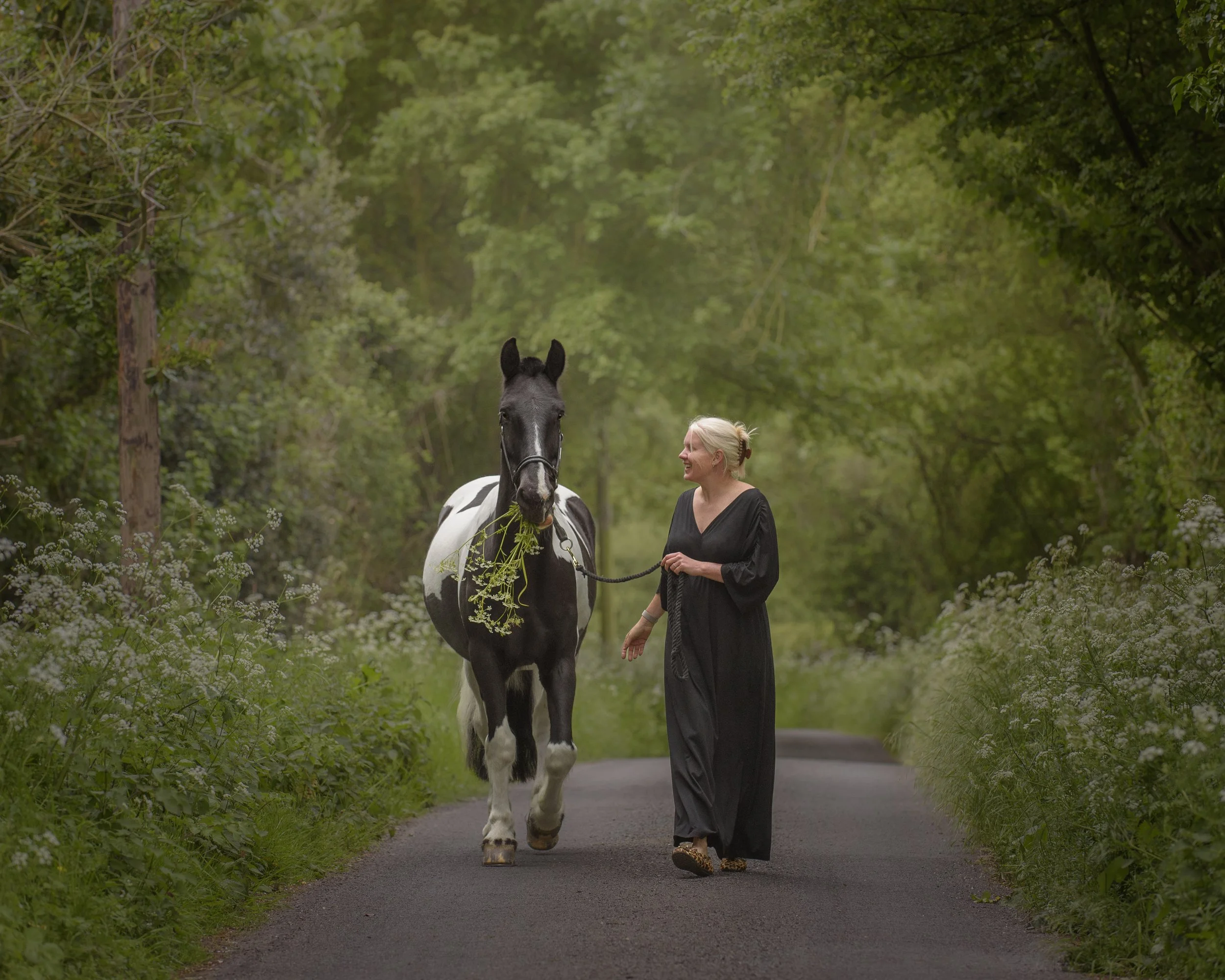 A woman with blonde hair in a bun walking next to a black and white horse along a paved path surrounded by green trees and bushes in Suffolk.