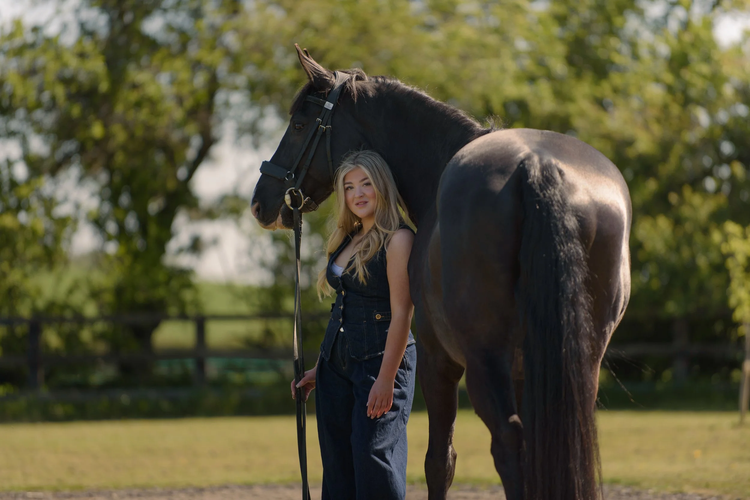Young woman standing next to a dark brown horse outdoors during daytime, surrounded by trees, with the horse's head near her shoulder and the woman smiling softly at the camera.
