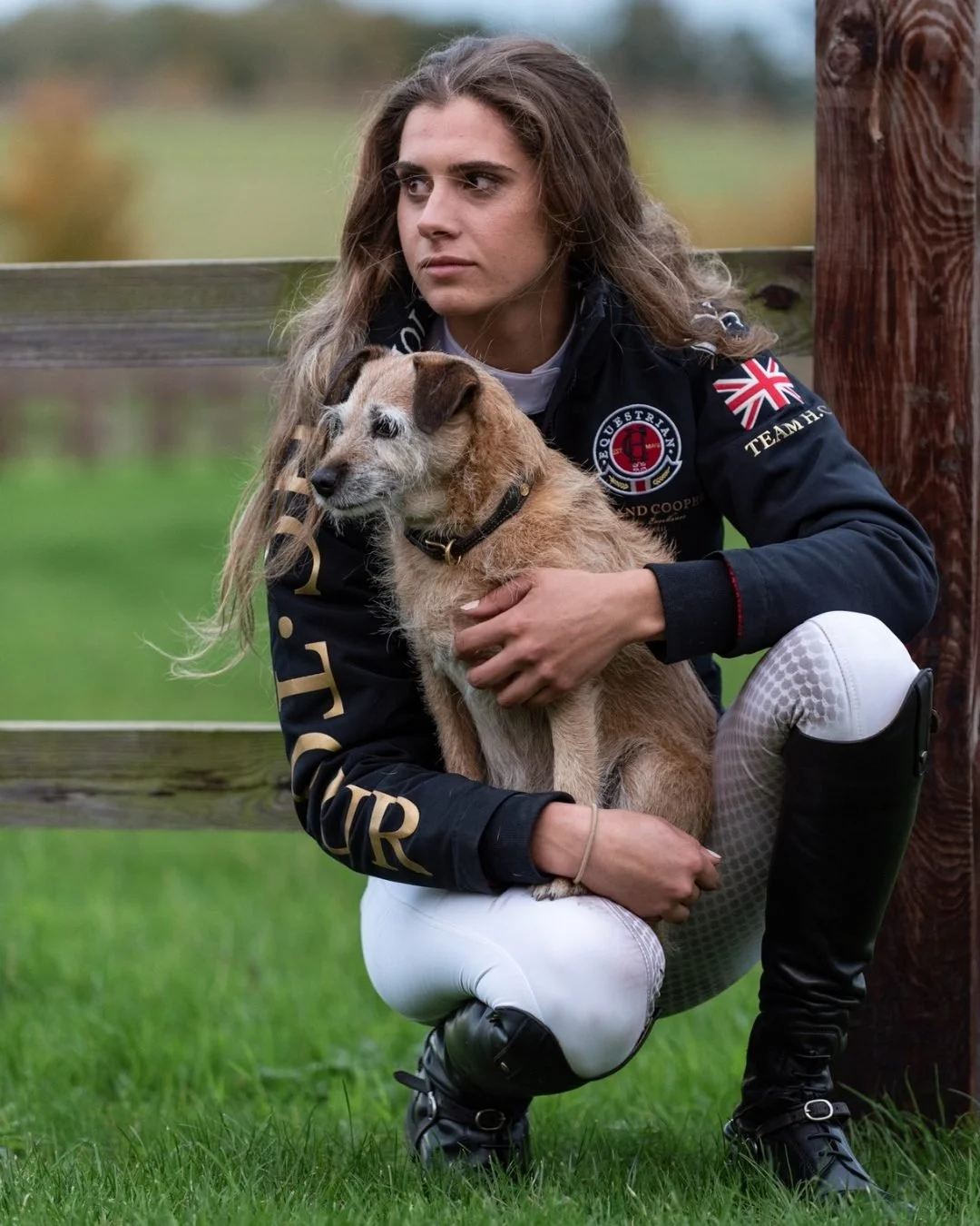 A woman with long hair crouches on grass, holding a small tan and white dog, with a wooden fence and blurred outdoor landscape in the background.