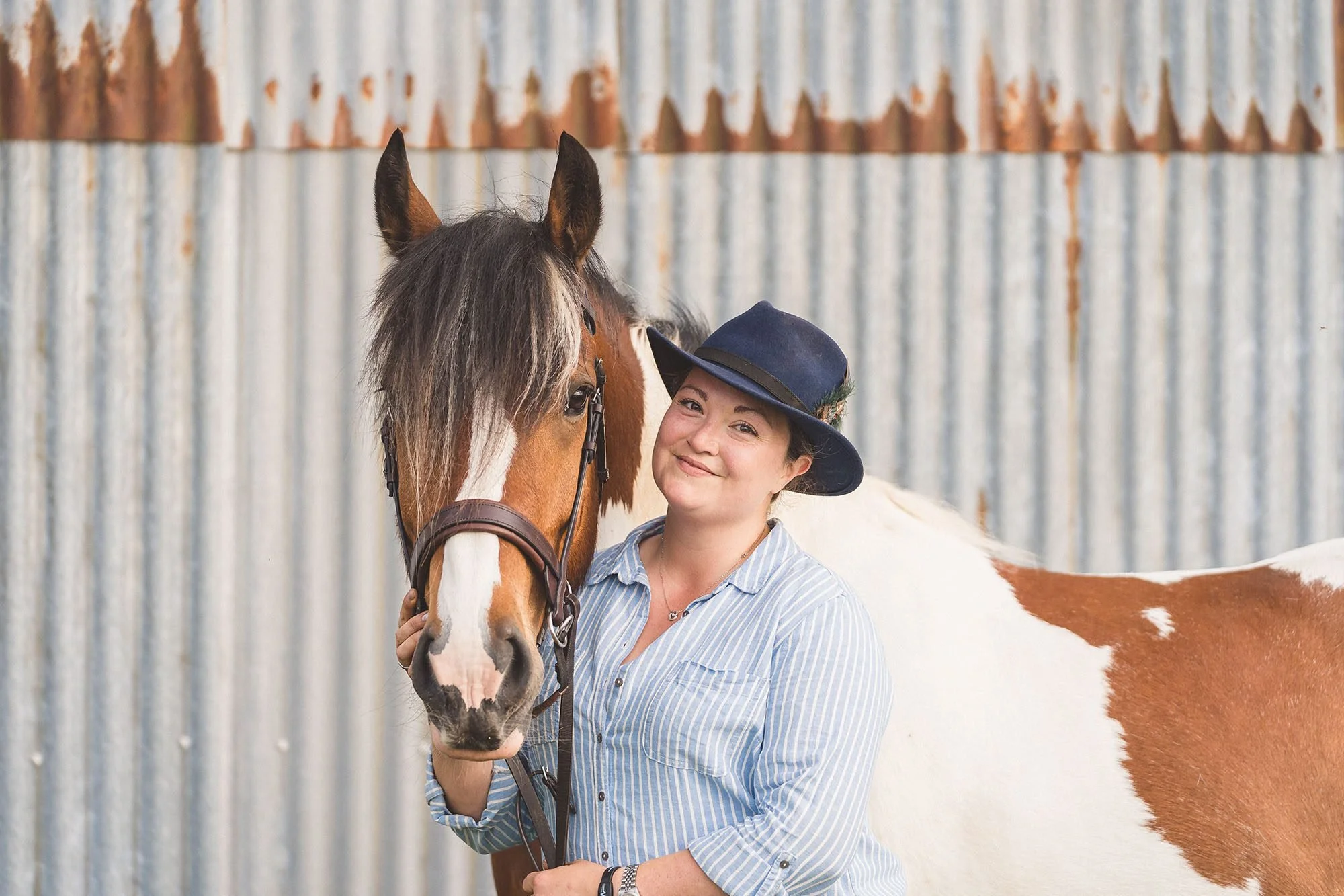 Young woman in a blue and white striped shirt and a navy hat standing next to a brown and white pinto horse with a brindle coat pattern, set against a backdrop of a corrugated metal wall.