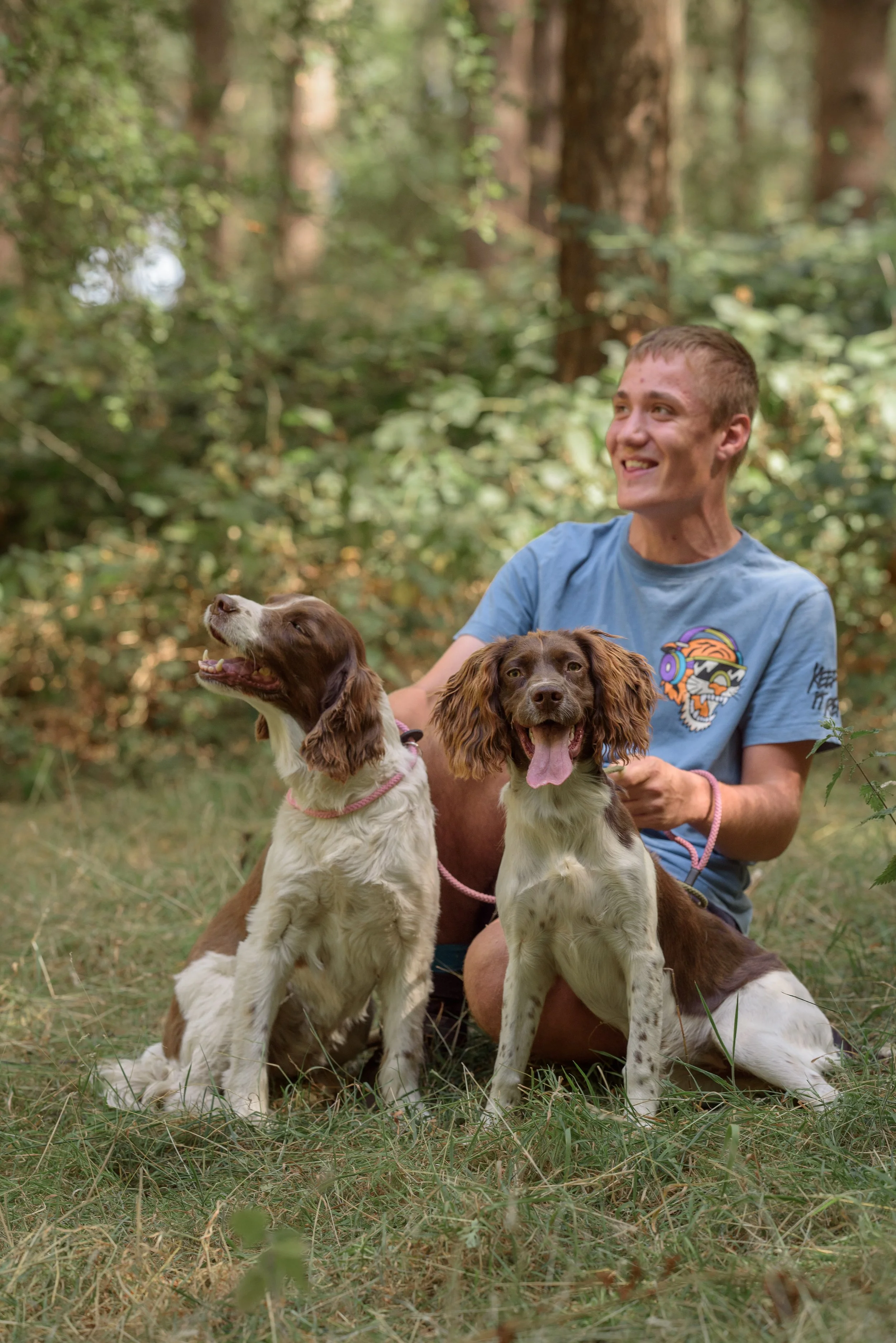 A young man in a blue T-shirt with a colorful tiger face on it, smiling and kneeling on the grass in a forest, holding two English Springer Spaniel dogs, one with its tongue out and the other looking up, with trees and greenery in the background.