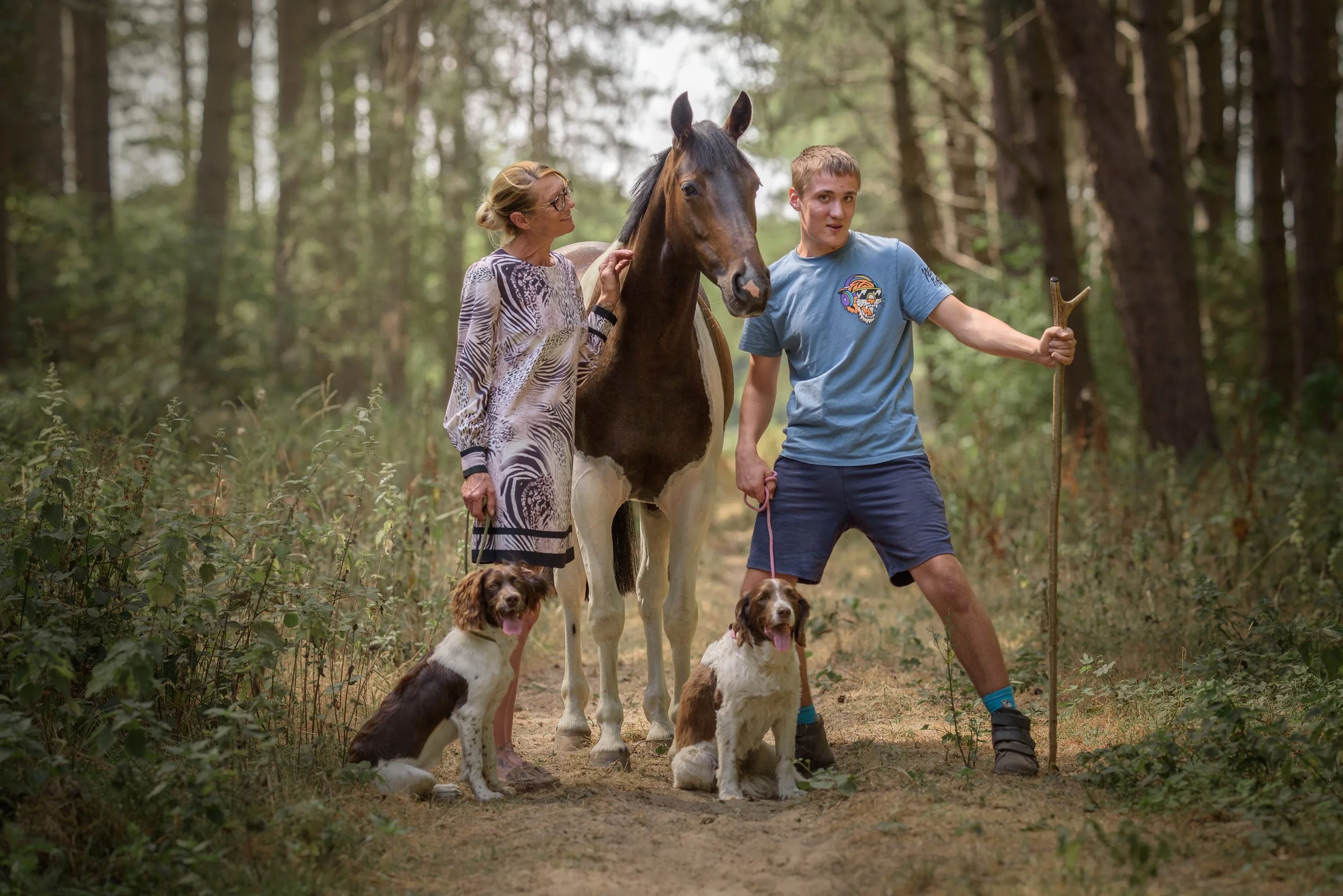 Two dogs sitting in front of a horse, with a mum and teenage son outdoors in a forest, holding the horse's reins and a walking stick.