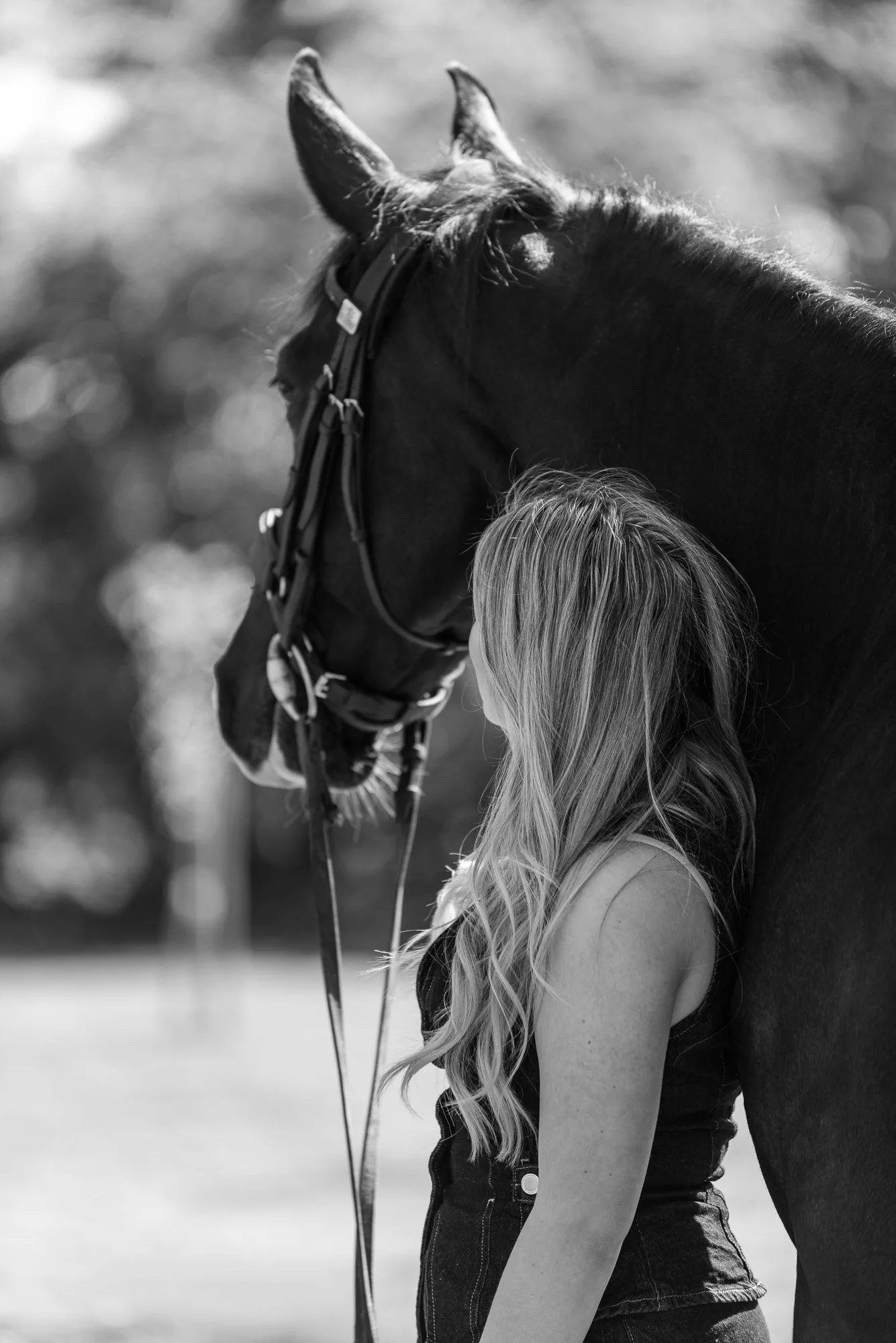 A young girl with long blonde hair standing close to a horse, both facing to the left. The girl is holding the horse's reins and appears to be gazing at it, with a blurred outdoor background.