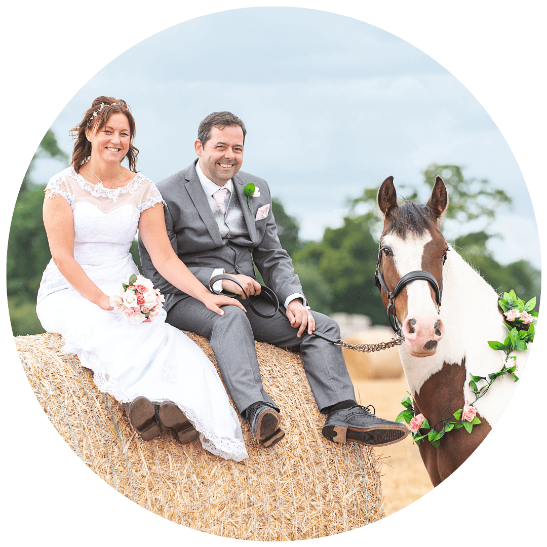 A bride and groom sitting on a hay bale in a field, smiling, with a horse decorated with flowers nearby, Essex.