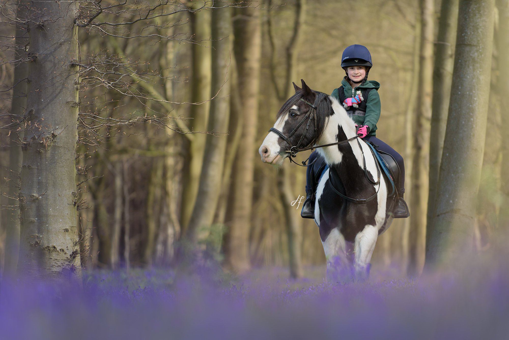 Young girl riding a black and white horse through a wooded area, wearing a helmet and green jacket.