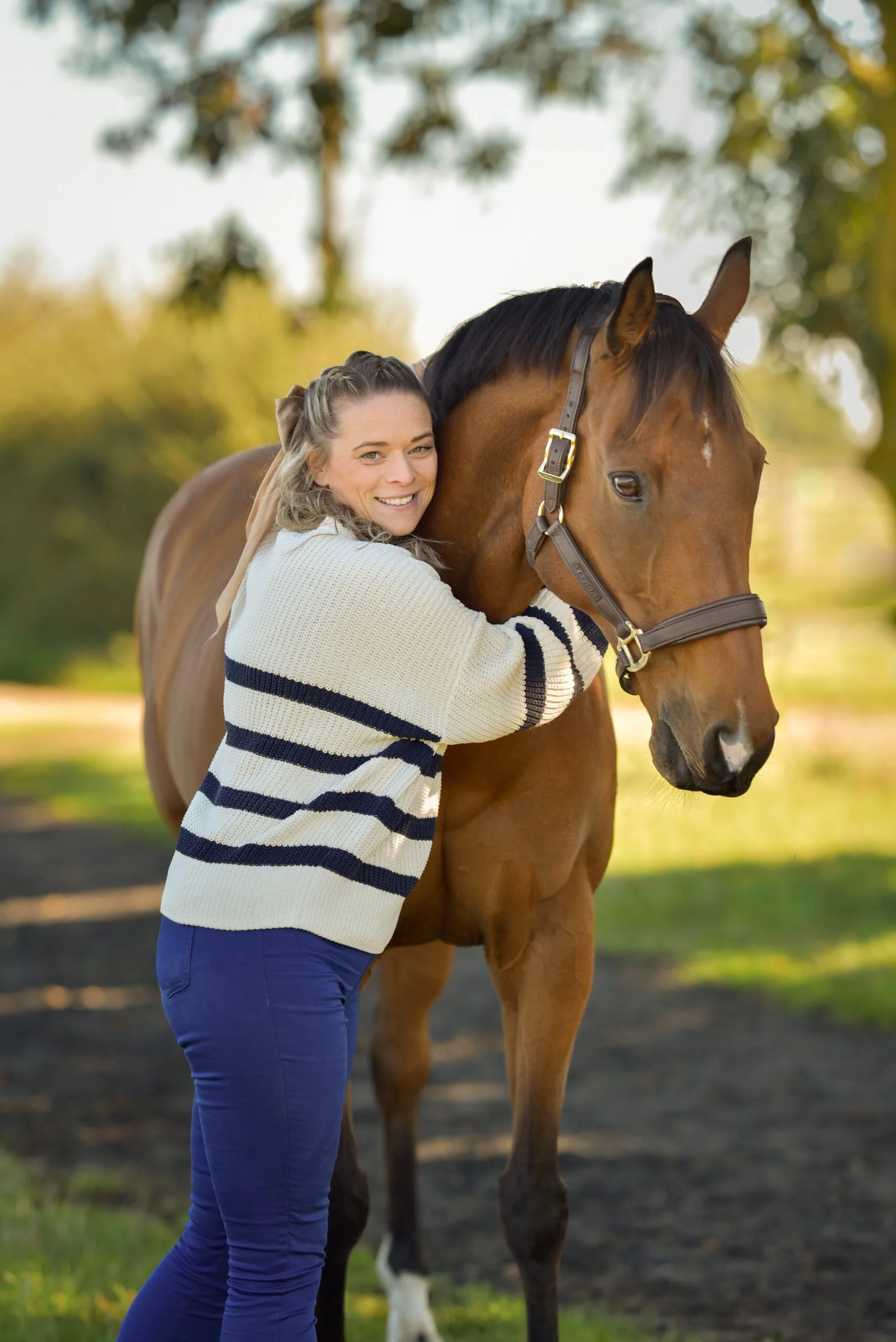 A woman hugging a brown horse outdoors with a background of blurred trees and sunlight.