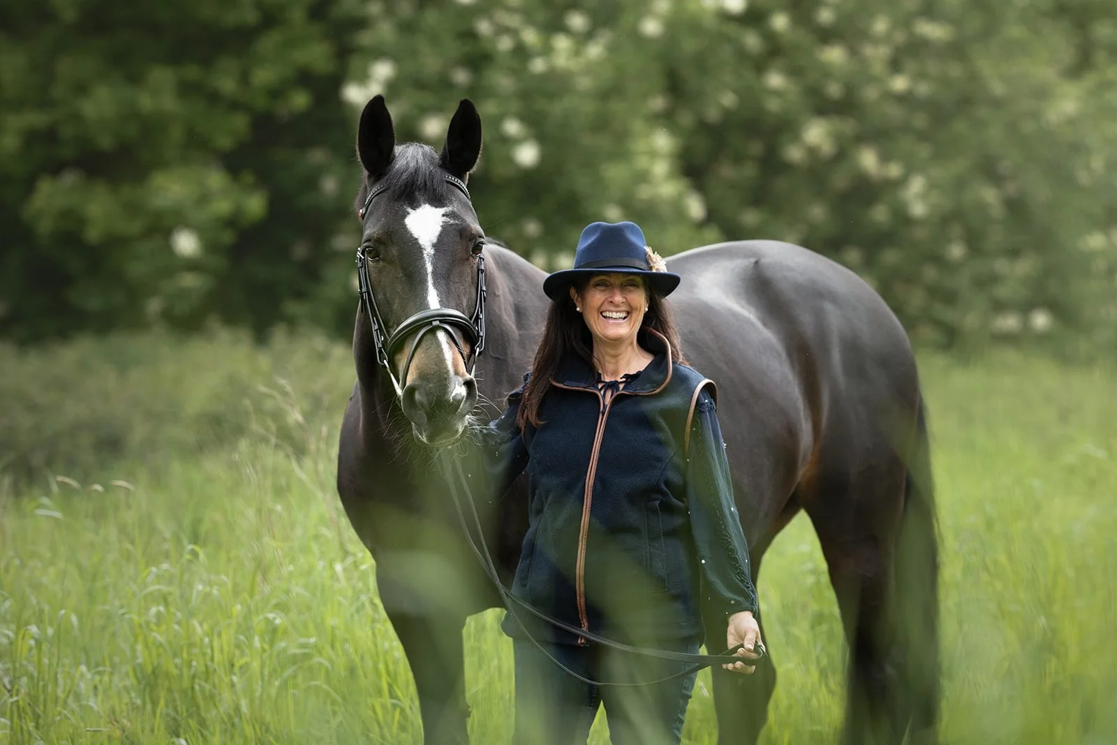 A woman smiling and walking alongside a large dark horse in a grassy field with trees in the background.