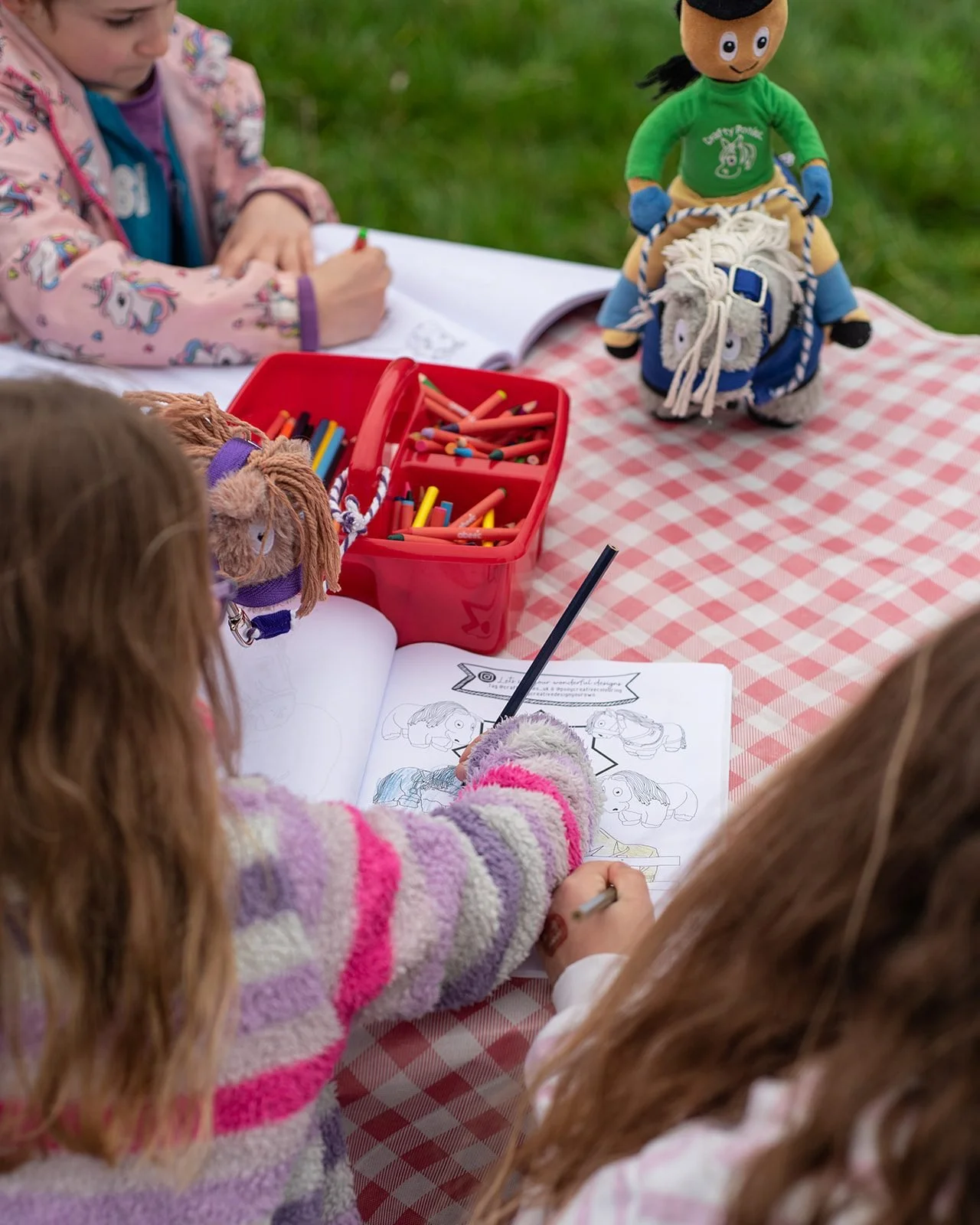 Children sitting at a picnic table outdoors, drawing and coloring with coloring books and crayons, with plush horse toys nearby.