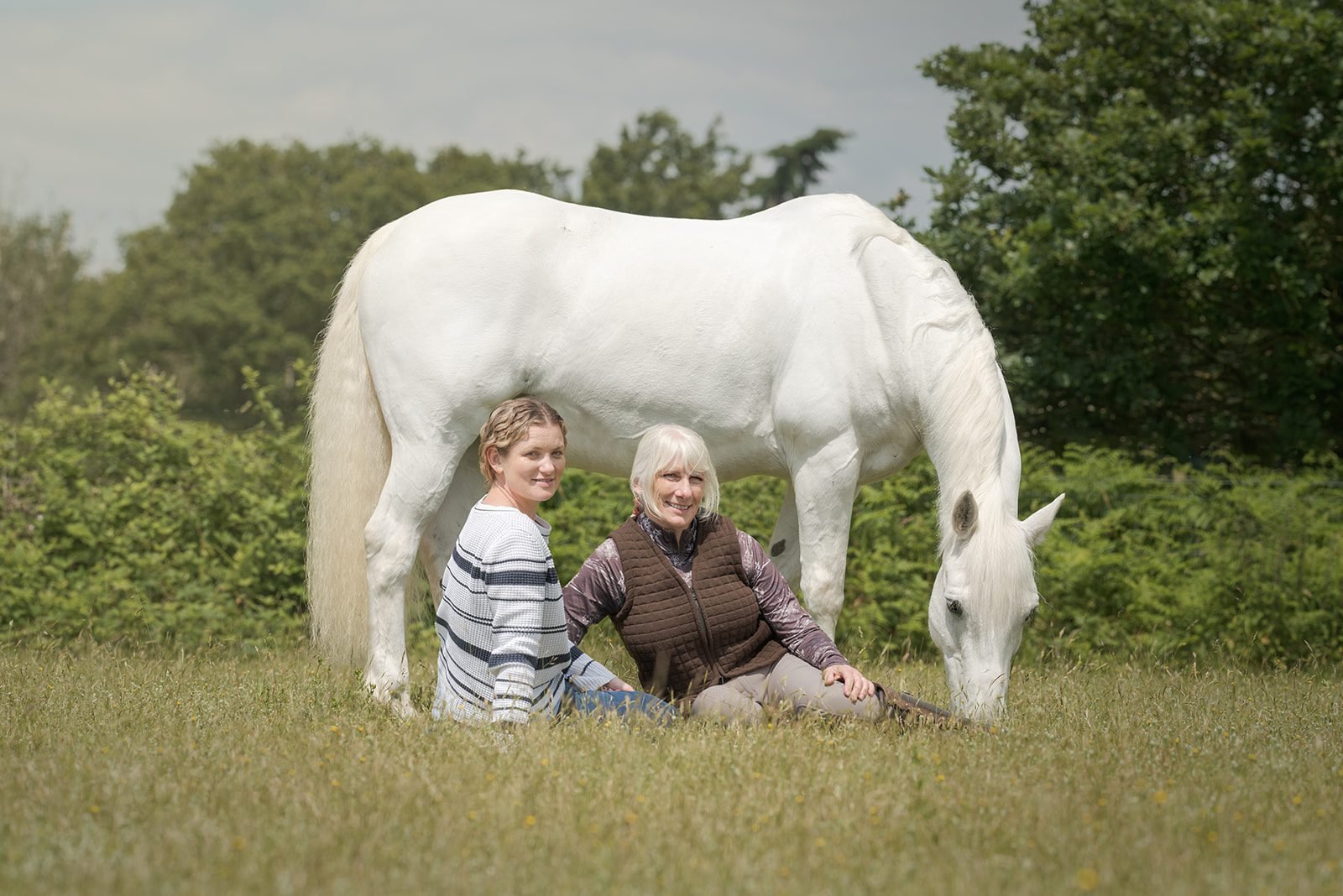 A woman with platinum blonde hair dressed in a long black dress is standing on a rural path, hugging a large black horse with a white star-shaped mark on its face. They are surrounded by lush green trees and foliage.