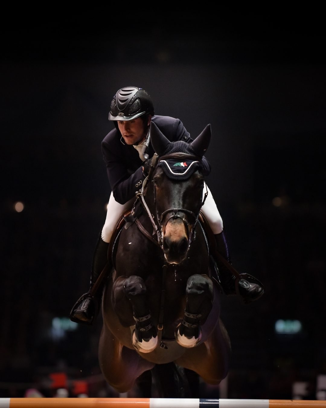 An equestrian athlete in riding attire and a helmet jumping over an obstacle on a dark horse during an indoor show jumping event.