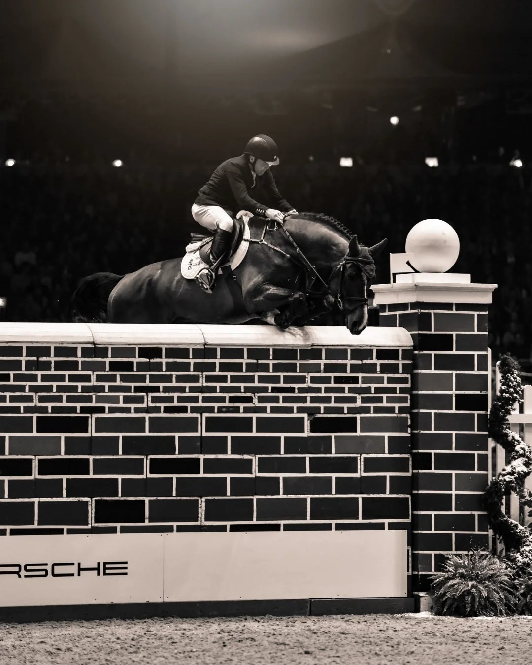 A horse and rider in a show jumping competition, mid-air over a brick obstacle in an indoor arena.