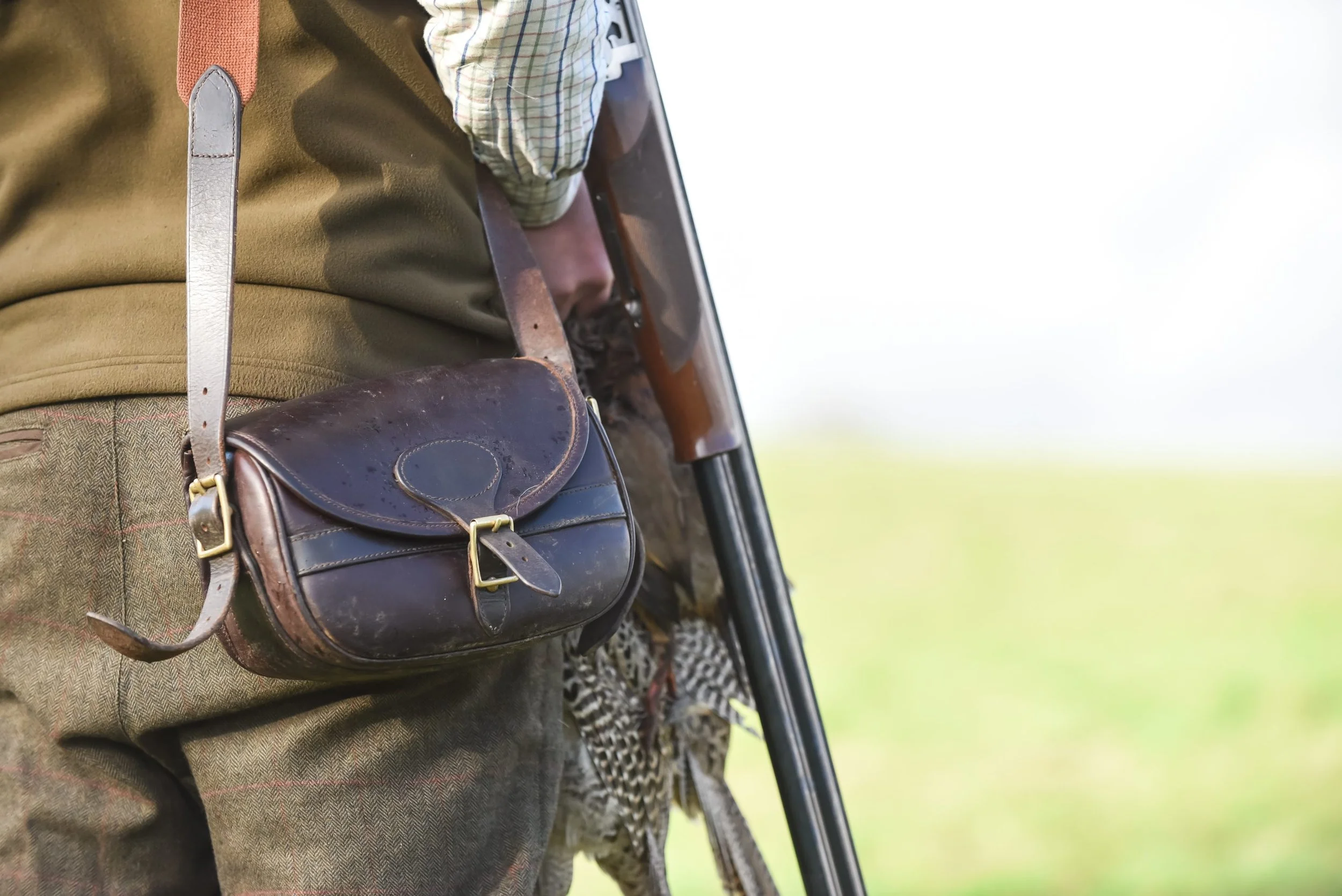 Close-up of a person wearing a plaid shirt and brown jacket, with a leather shoulder bag hanging from their shoulder, holding a shotgun and grouse, outdoors on a grassy background.