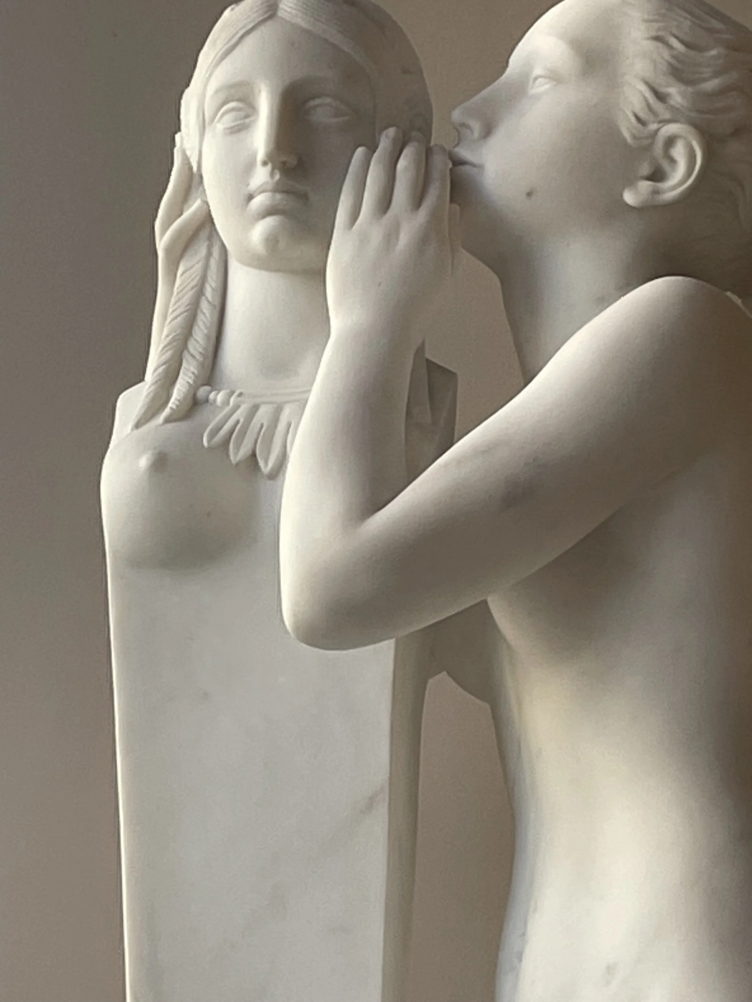 Close-up of a white marble sculpture depicting a man and a woman in a tender moment, with the woman whispering into the man's ear.