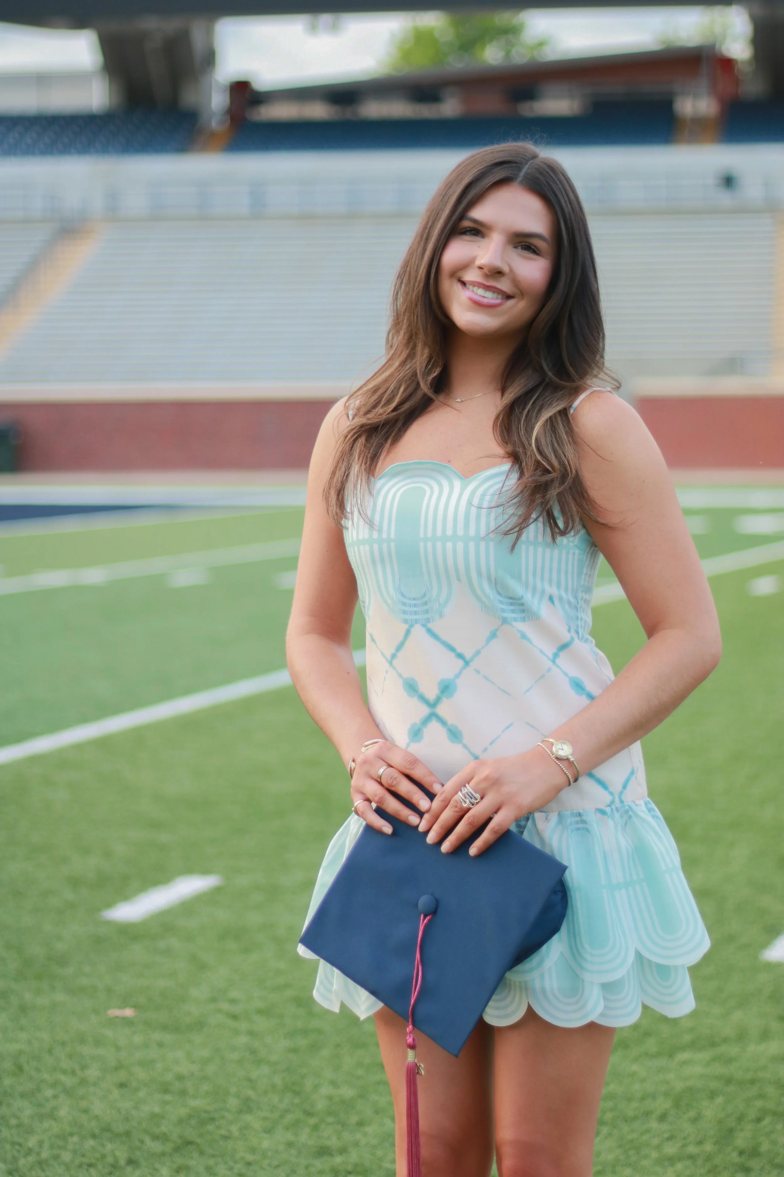 A young woman with long brown hair, smiling, wearing a light blue and white patterned dress, holding a navy blue graduation cap, standing on a football field.