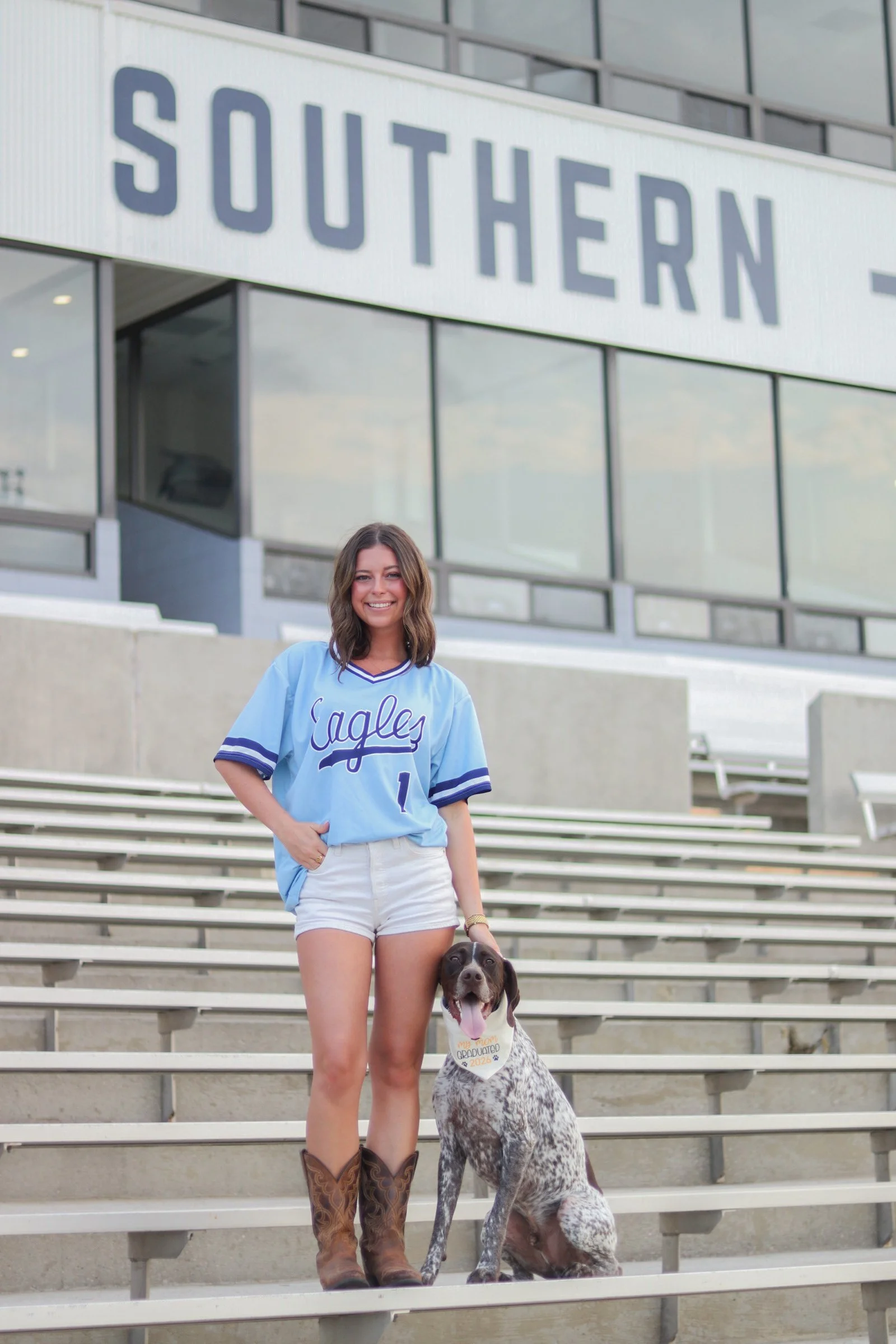 A young woman in a light blue sports jersey and white shorts stands on stadium bleachers, smiling at the camera. She has brown hair and is holding a large, spotted dog that sits beside her with a graduation cap on its collar. The background features 