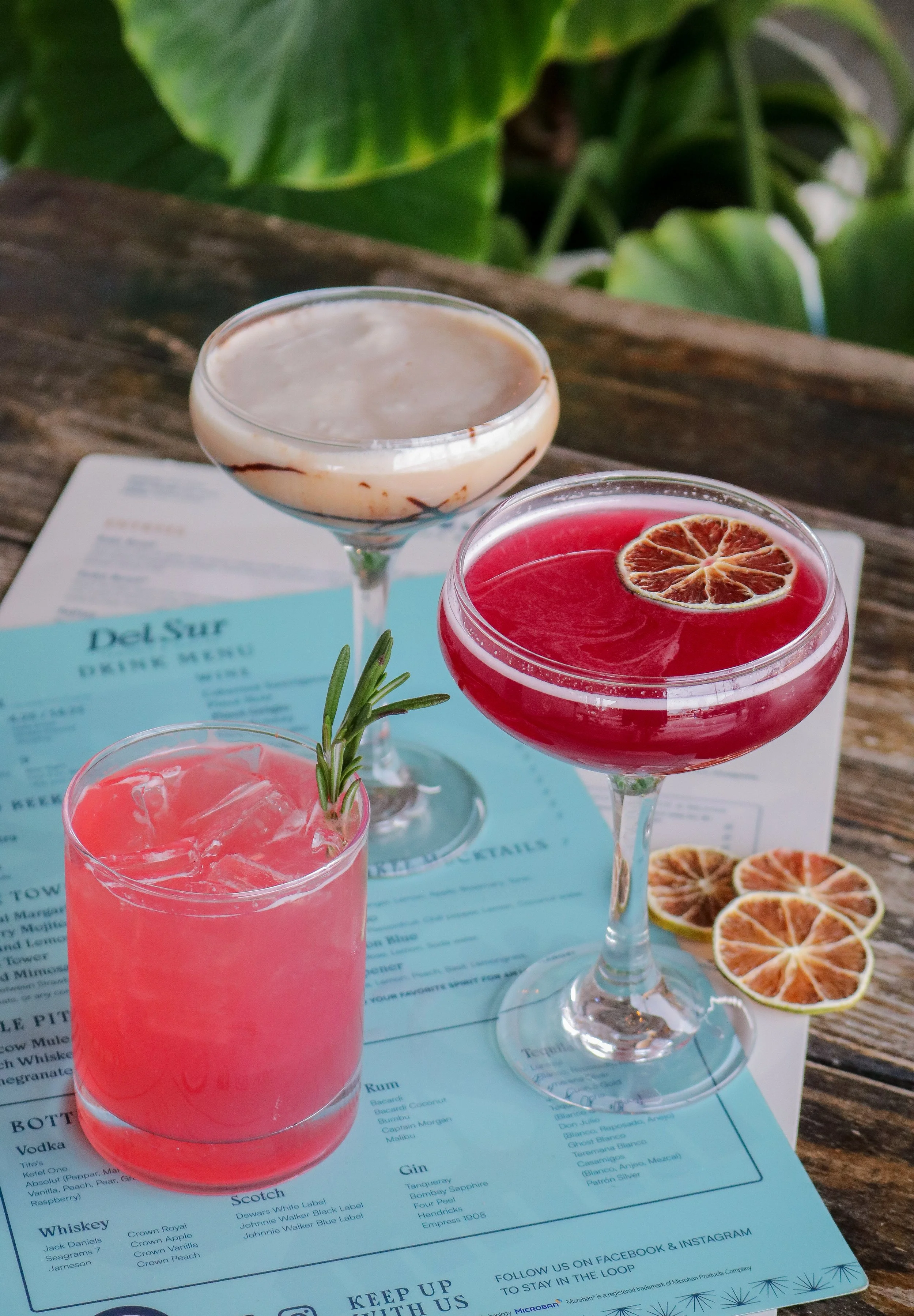 Three colorful cocktails on a wooden table with a blue drink menu underneath. The drinks include a red drink with a dried citrus slice, a pink drink with a sprig of rosemary, and a creamy drink with a chocolate drizzle.