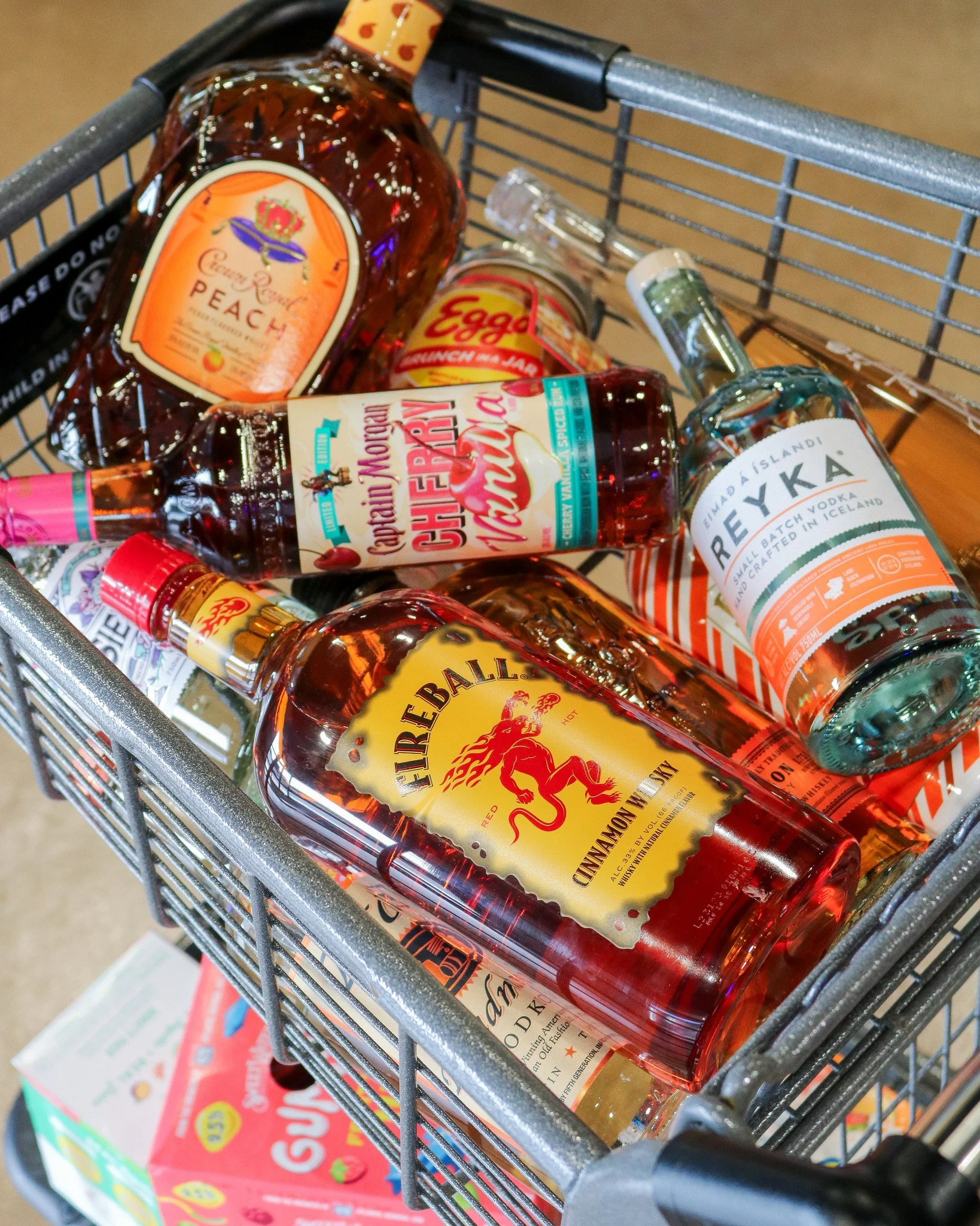 A shopping cart filled with various bottles of alcohol, including Fireball cinnamon whisky, Bæyka vodka, and a peach-flavored aperitif, in a store aisle.