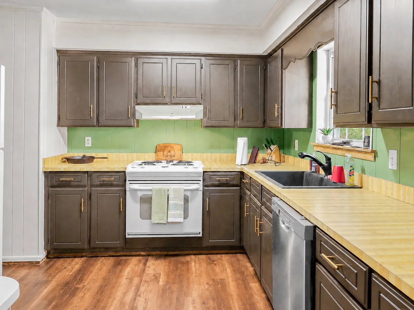 Kitchen with dark gray cabinets, yellow wooden countertops, a white stove, stainless steel dishwasher, black sink, green backsplash, small window with plants, and hardwood floor.