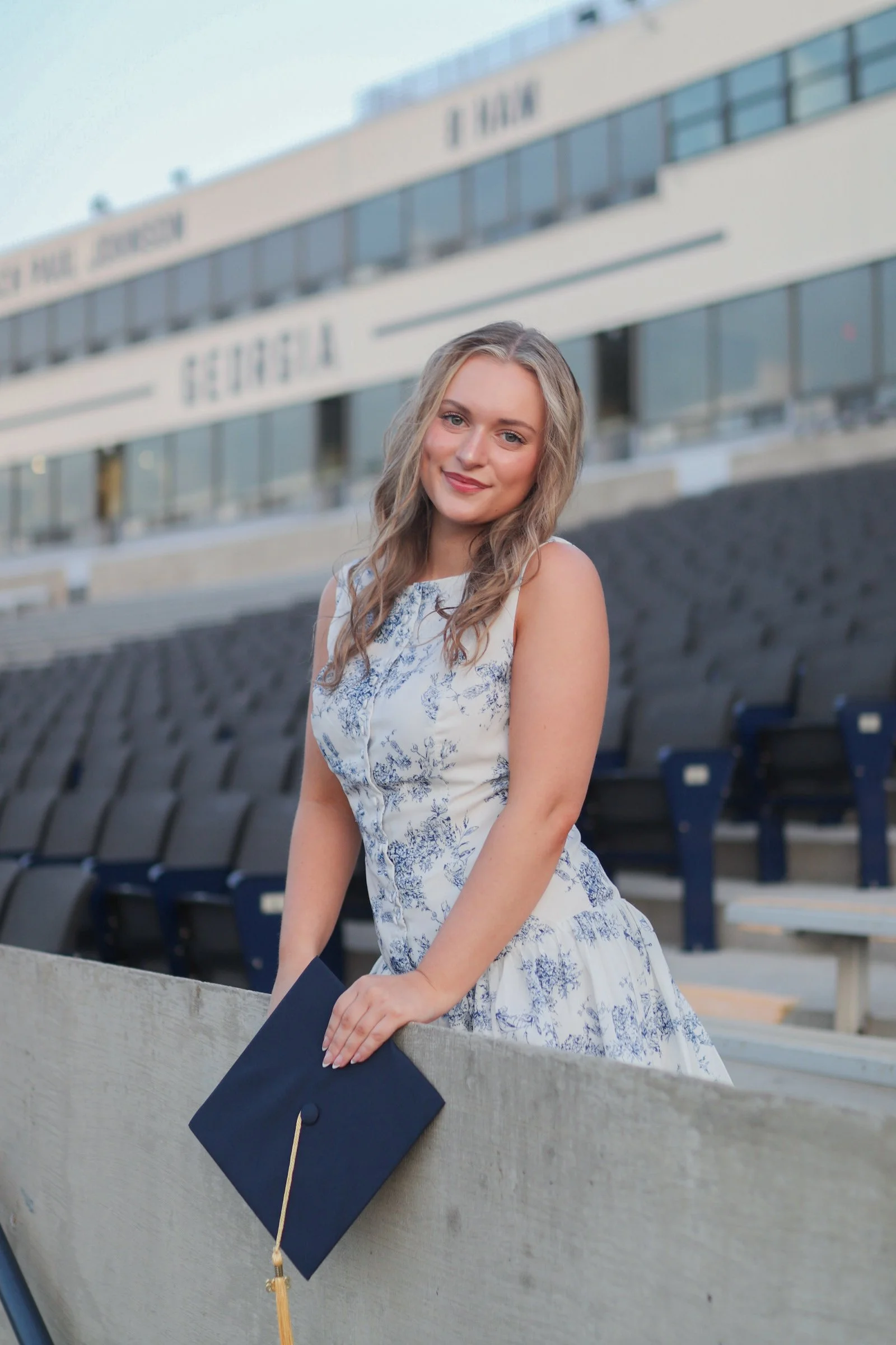 A young woman in a white and blue floral dress holding a diploma in front of a stadium stands, celebrating graduation day.