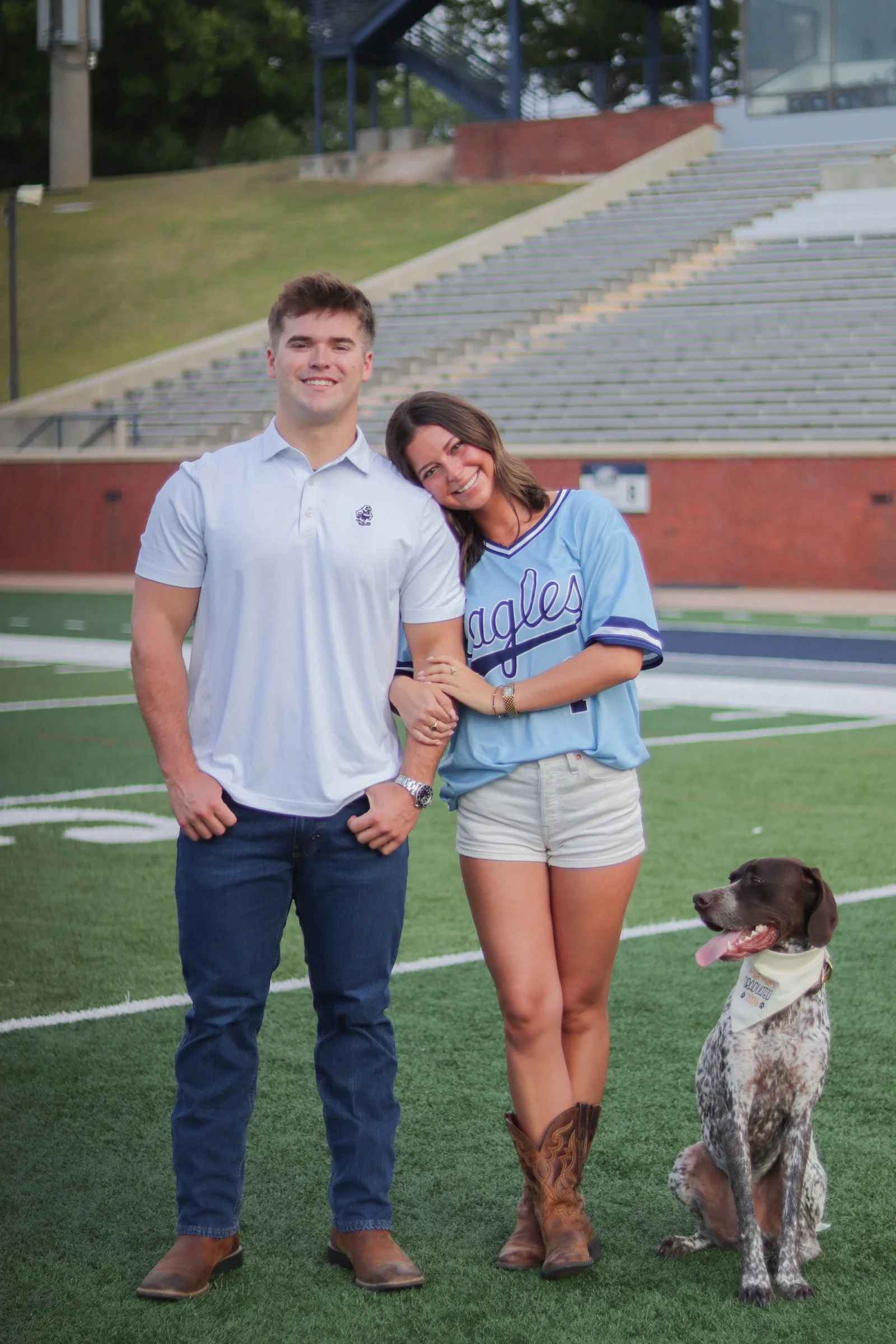 A smiling man and woman stand on a football field with a dog sitting beside them. The man is wearing a white polo shirt and jeans, the woman is in a blue sports jersey and shorts, and the dog is a German Shorthaired Pointer wearing a bandana. The bac