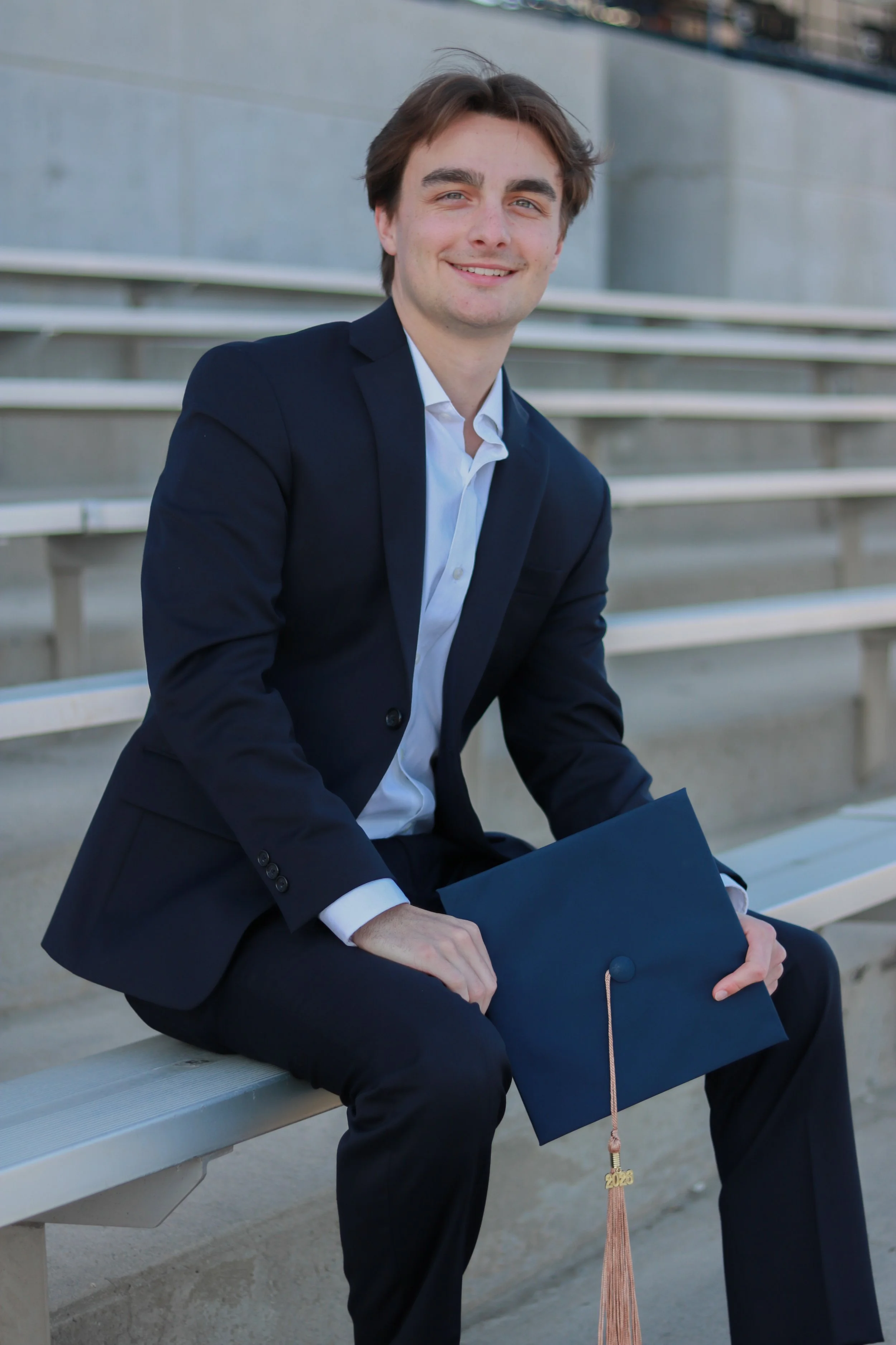 A young man in a dark suit sitting on outdoor bleachers, holding a graduation cap with a tassel and a year charm that says 2020.