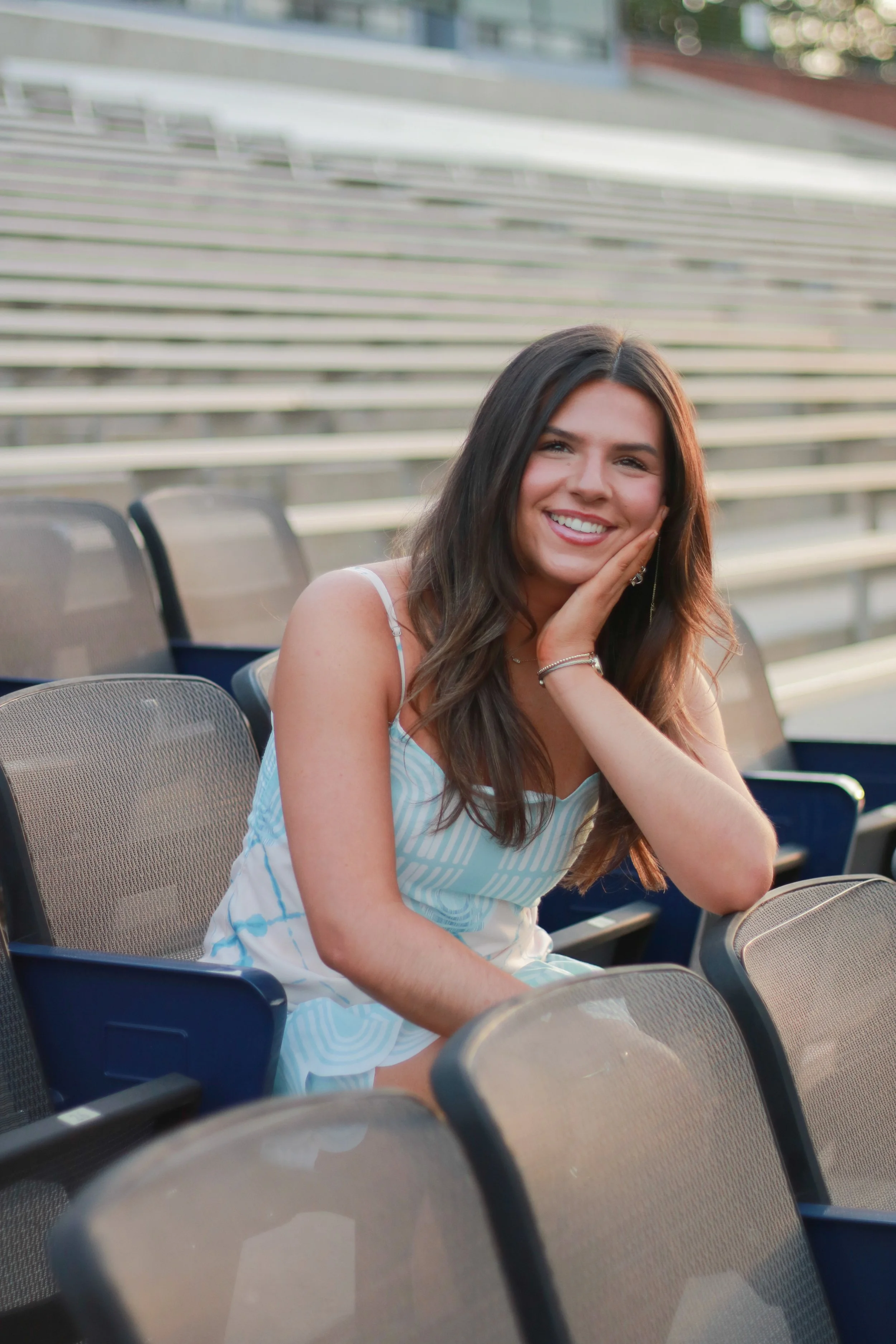 A young woman with long, dark hair smiling, sitting outdoors in a stadium with empty seats in the background, wearing a white and light blue striped sleeveless dress.
