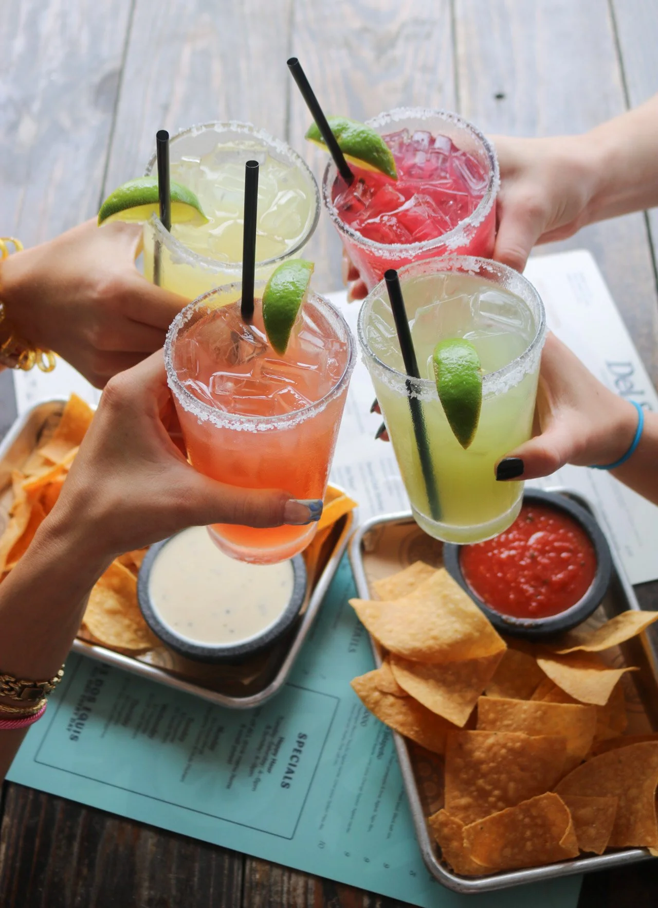 Four hands holding colorful cocktails with lime wedges, surrounded by chips, salsa, and creamy dips on a restaurant table.