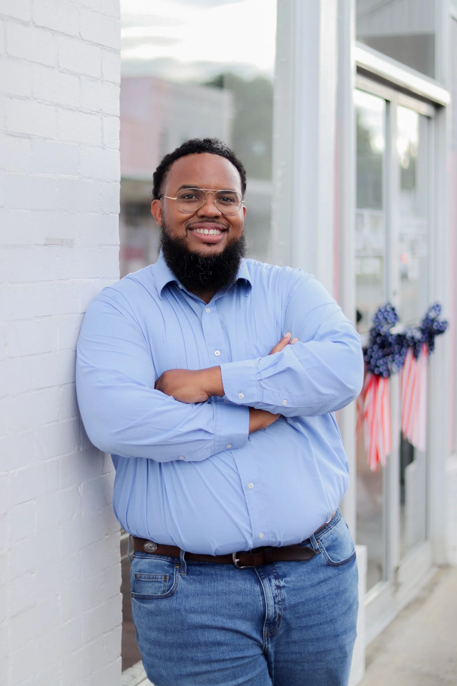 A man with glasses, a beard, wearing a light blue button-up shirt and jeans, standing with arms crossed and smiling against a white brick wall outside.