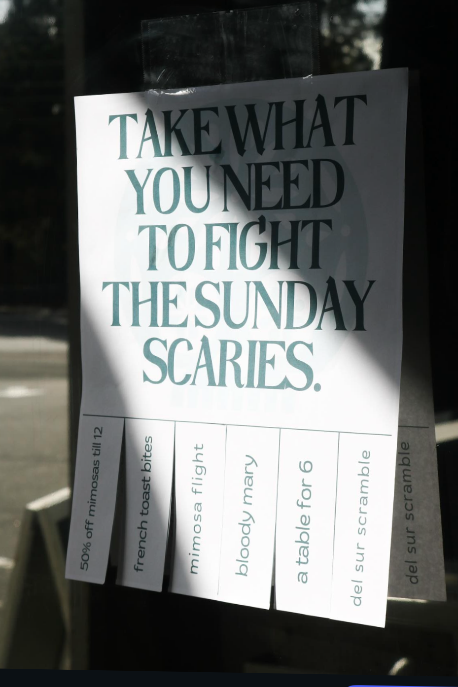 A sign on a window with the phrase "Take what you need to fight the Sunday scaries"; below, there are small notes with various phrases, such as "50% off mimosas till 12," "French toast bites," "mimosa flight," "bloody mary," "a table for 6," and "del